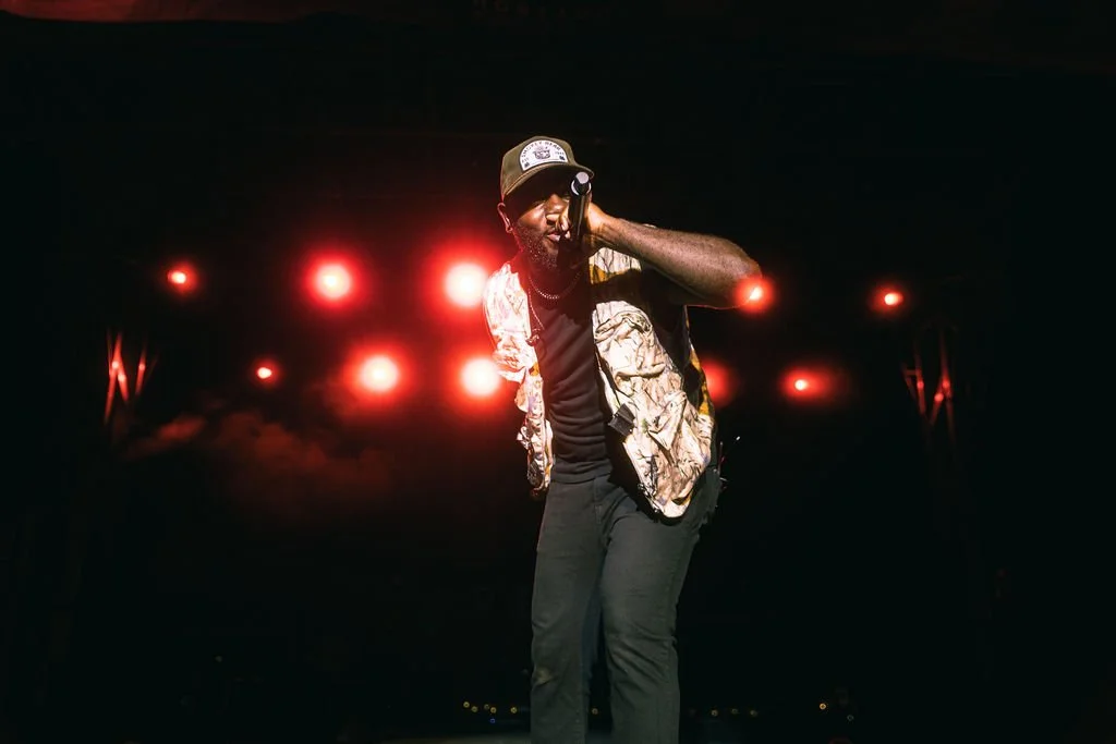 A male performer singing into a microphone on stage with bright lights behind him.