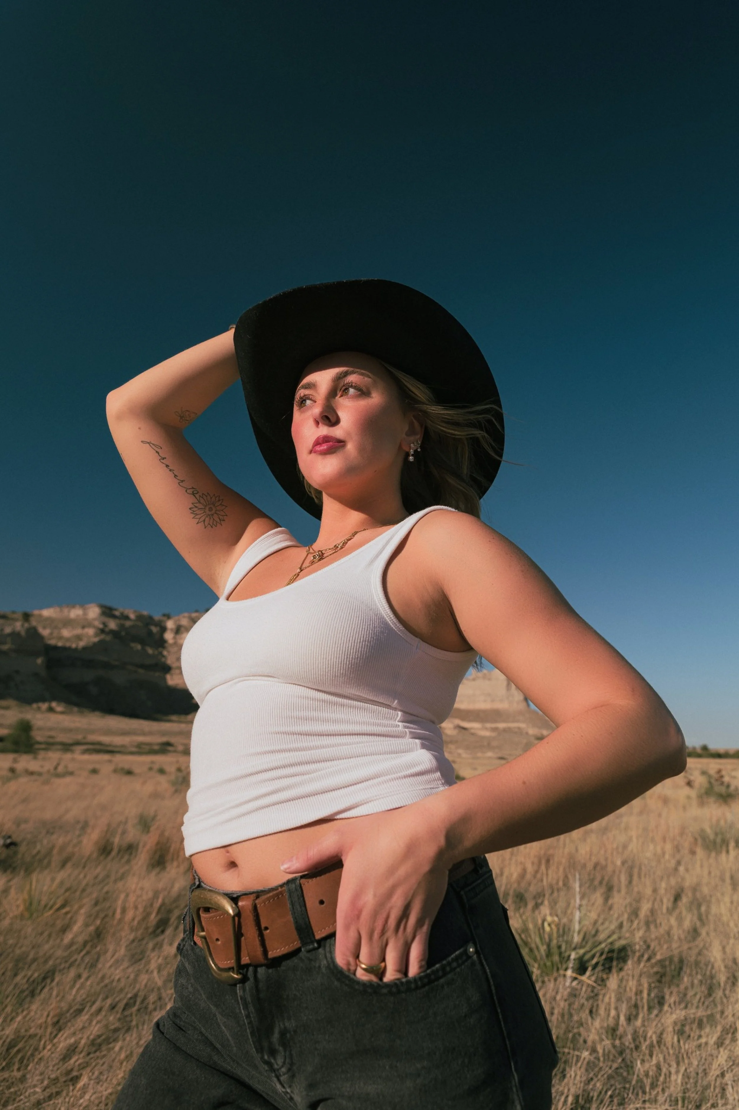 A woman wearing a wide-brimmed black hat, white tank top, and dark jeans stands outdoors in a dry, grassy landscape. She is looking slightly upwards with her left hand resting on her head and her right hand on her waist. The sky is clear and blue, and distant hills are visible in the background.