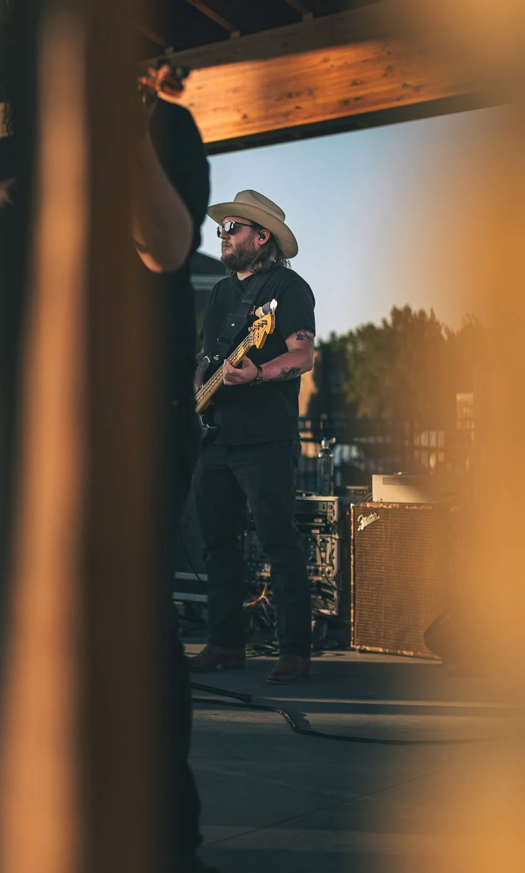 A musician wearing sunglasses, a wide-brimmed hat, and black clothing playing an electric guitar on stage during an outdoor concert at sunset; equipment and trees visible in the background.
