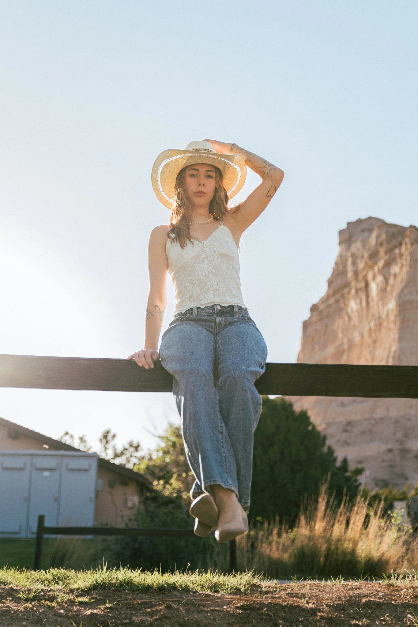 A young woman wearing a sunhat and casual clothes sitting on a wooden beam outdoors with a rocky hill in the background during sunset