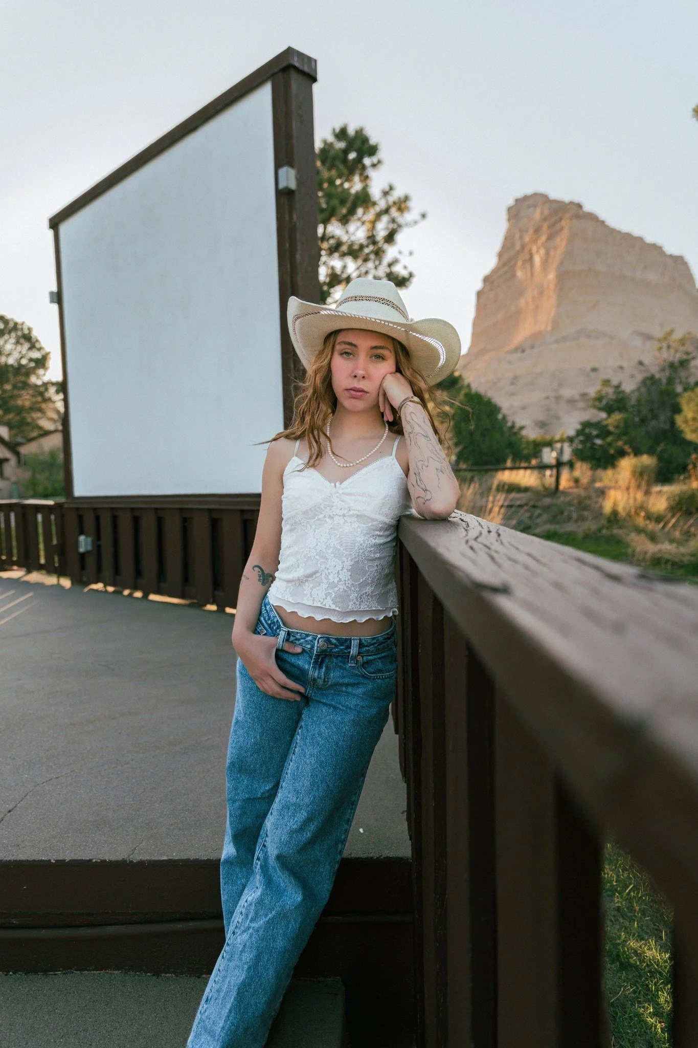 Young woman with wavy hair wearing a white lace camisole and blue jeans, leaning on a wooden railing with a desert-like mountain in the background, during early evening.