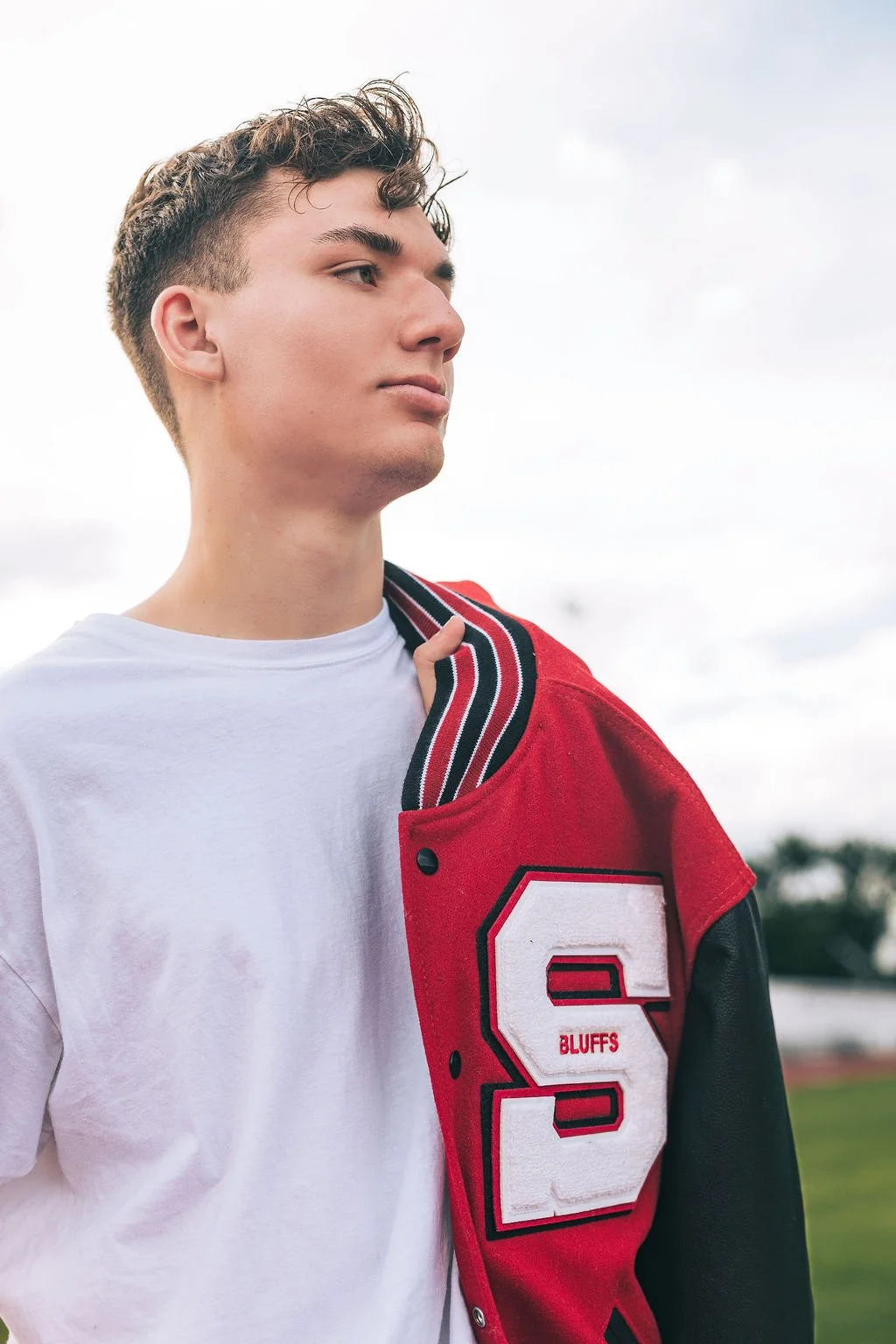 Young man outdoors wearing a red and black varsity jacket over a white t-shirt, looking to the side with a calm expression.