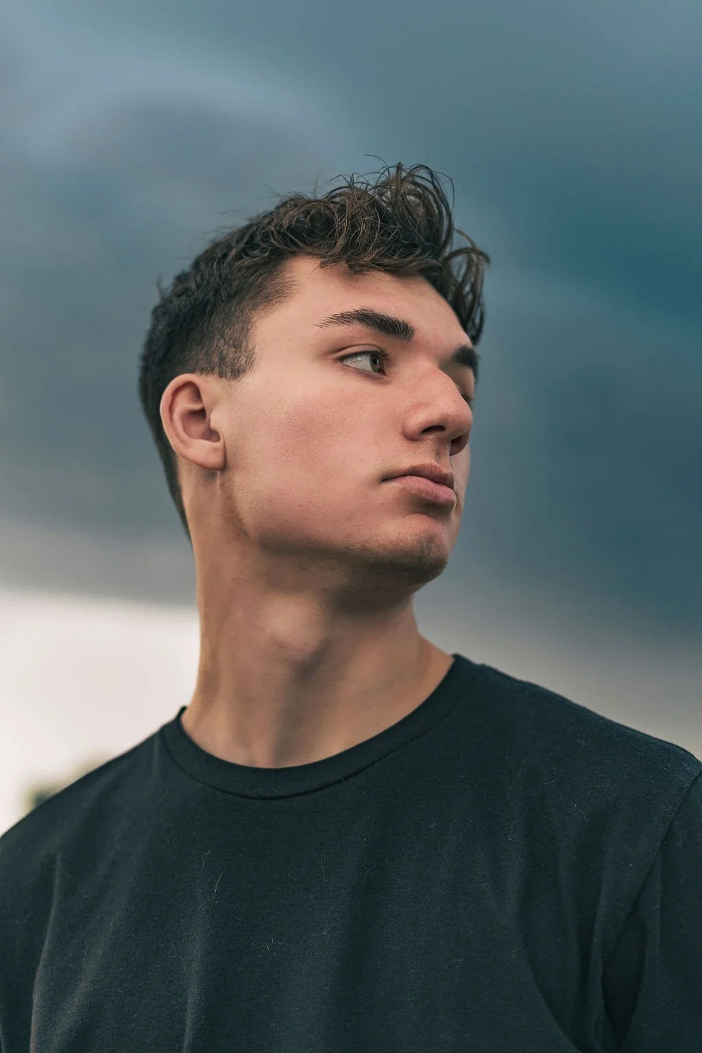Portrait of a young man with short brown hair, wearing a black shirt, looking to the side against a cloudy sky background.
