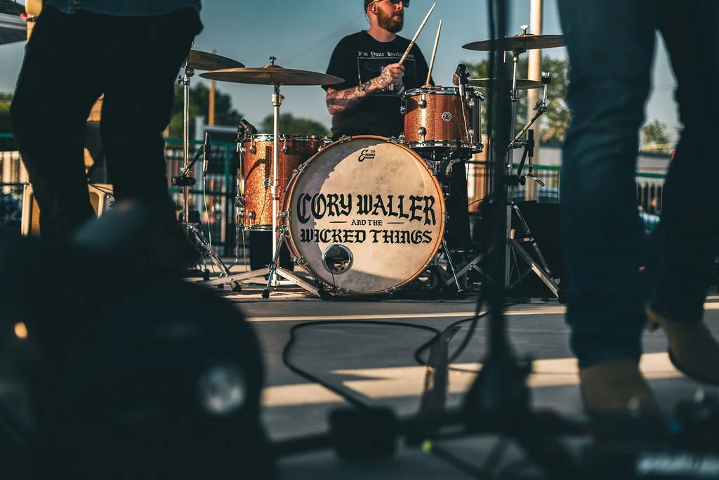 A drummer playing on a drum set with a bass drum that has the band name "Cory Waller and The Wicked Things" written on it, during an outdoor performance at sunset.