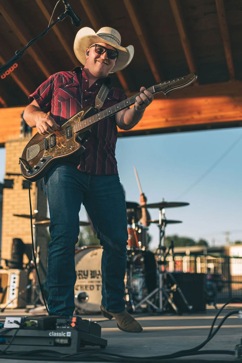 A man wearing a cowboy hat, sunglasses, and a striped shirt playing an electric guitar on an outdoor stage, with a drummer visible in the background.