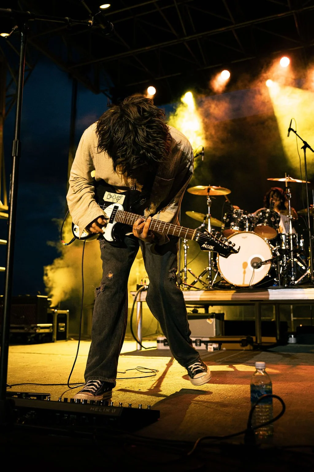 A guitarist performing on stage with a drummer in the background, colorful stage lights, and musical equipment, including a water bottle on the floor.