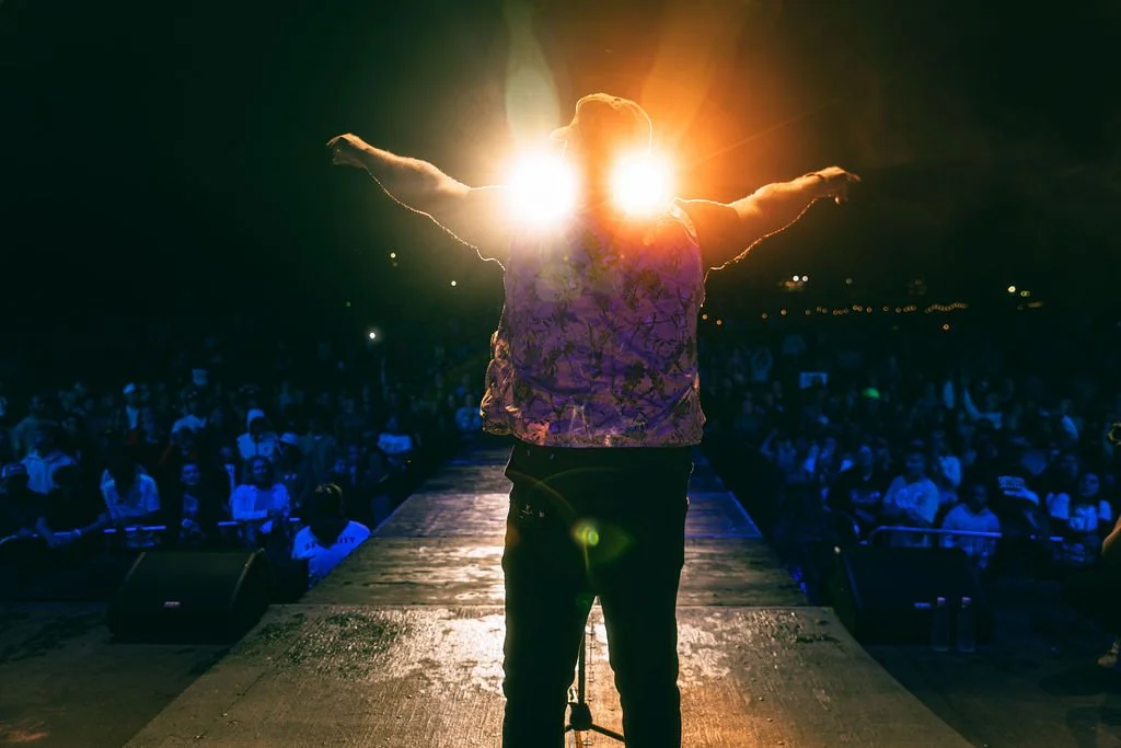 Performer on stage with arms outstretched, backlit by bright lights, facing a large audience at night.