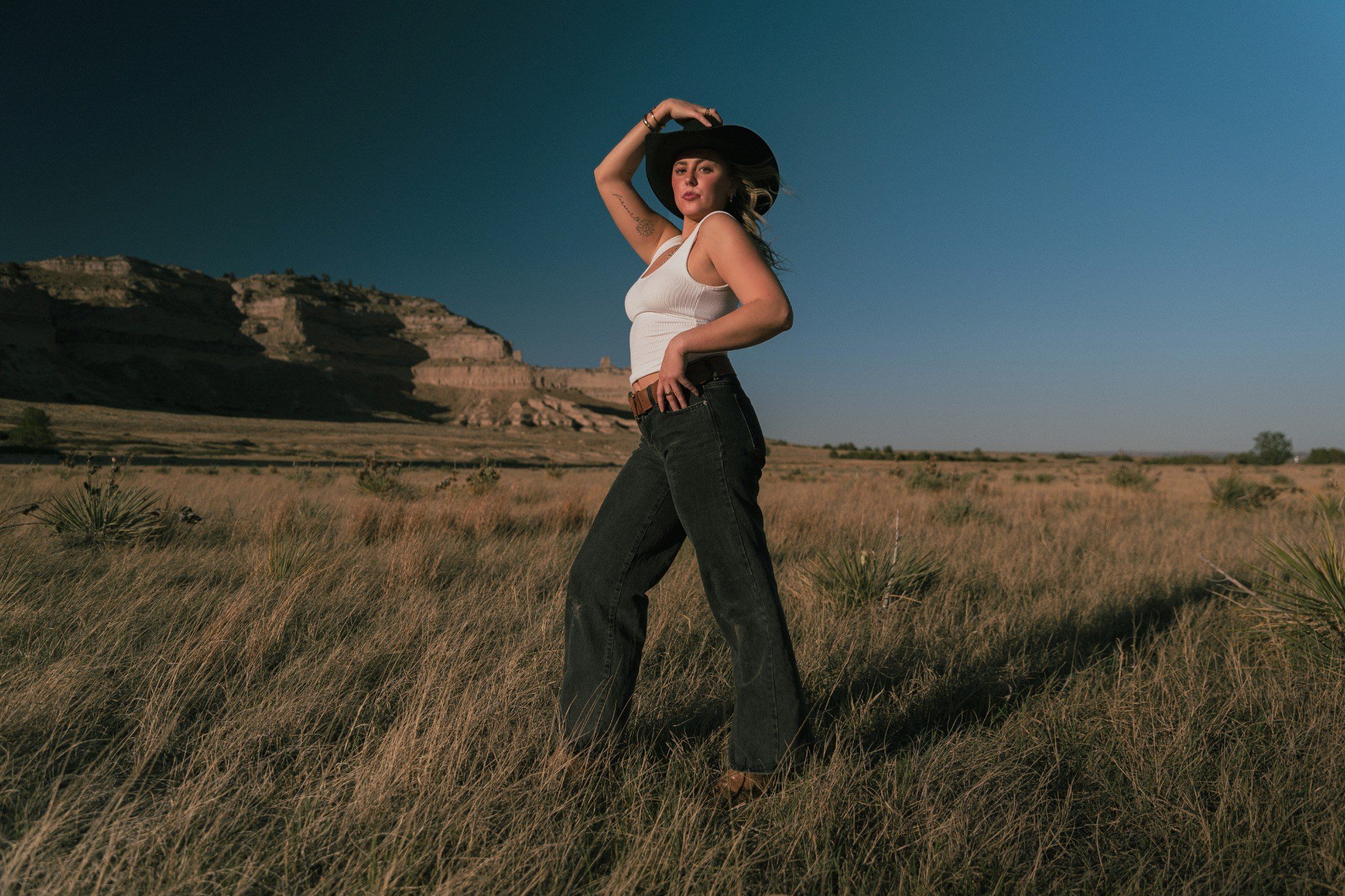 A woman in a white tank top and black jeans, wearing a wide-brimmed black hat, stands in a grassy plain with a rocky hill in the background, holding the hat with one hand and looking at the camera.