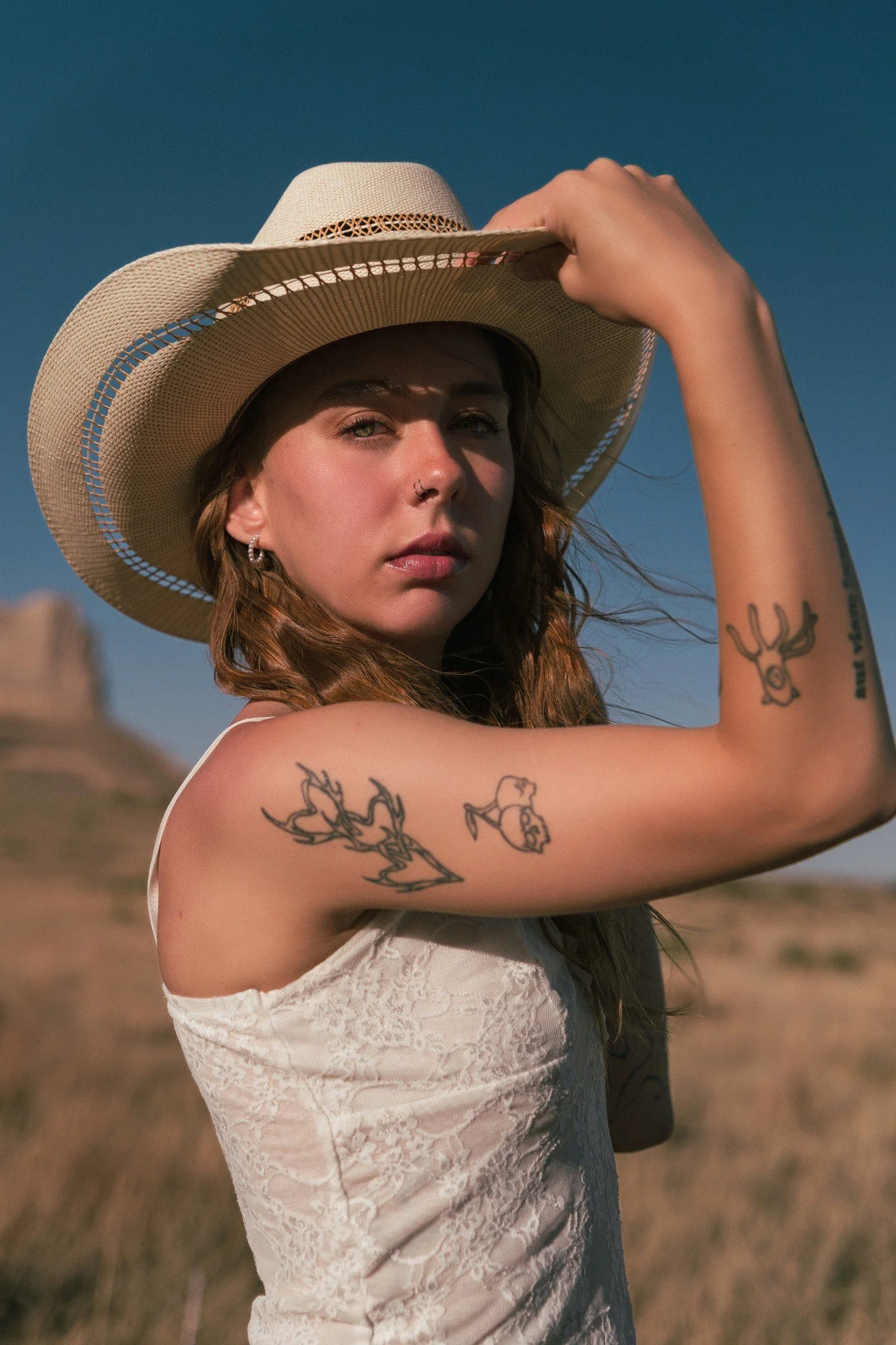 A woman with tattoos on her arm and earrings, wearing a lace dress and a large sun hat, outdoors with a clear sky background.