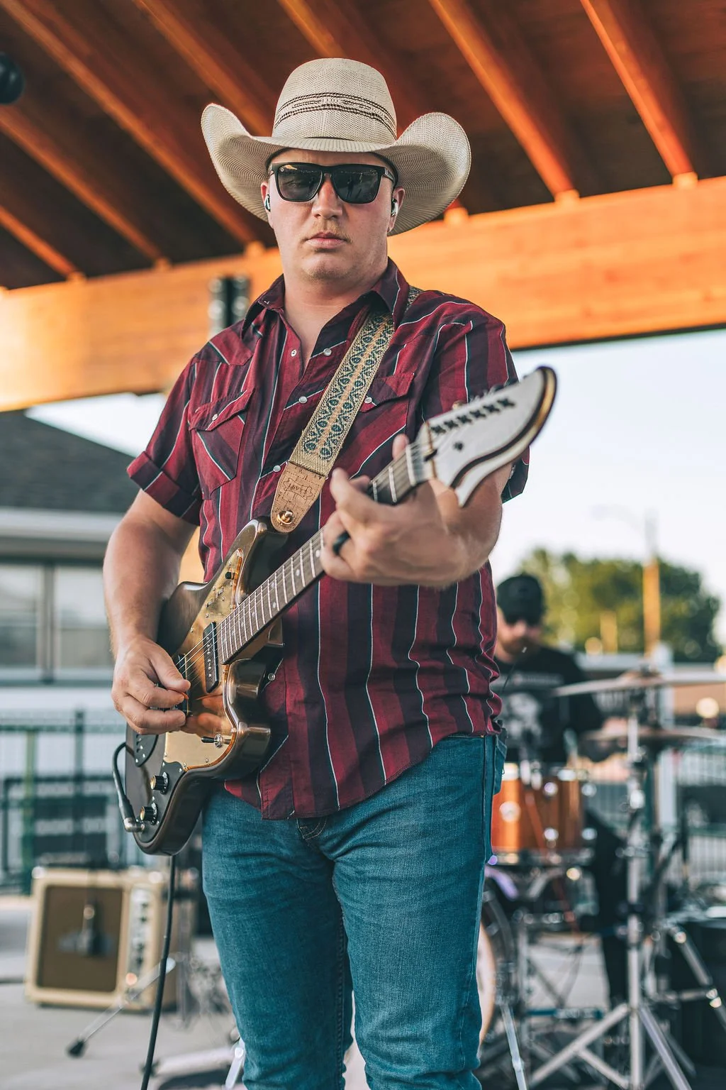 Man wearing a cowboy hat, sunglasses, and a red striped shirt playing an electric guitar outdoors.