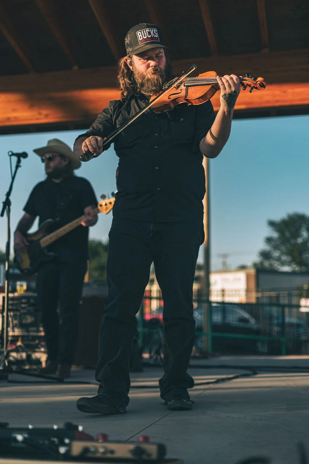 A man with a beard and long hair playing a violin on an outdoor stage, wearing a cap and black shirt, with another musician playing bass guitar in the background.