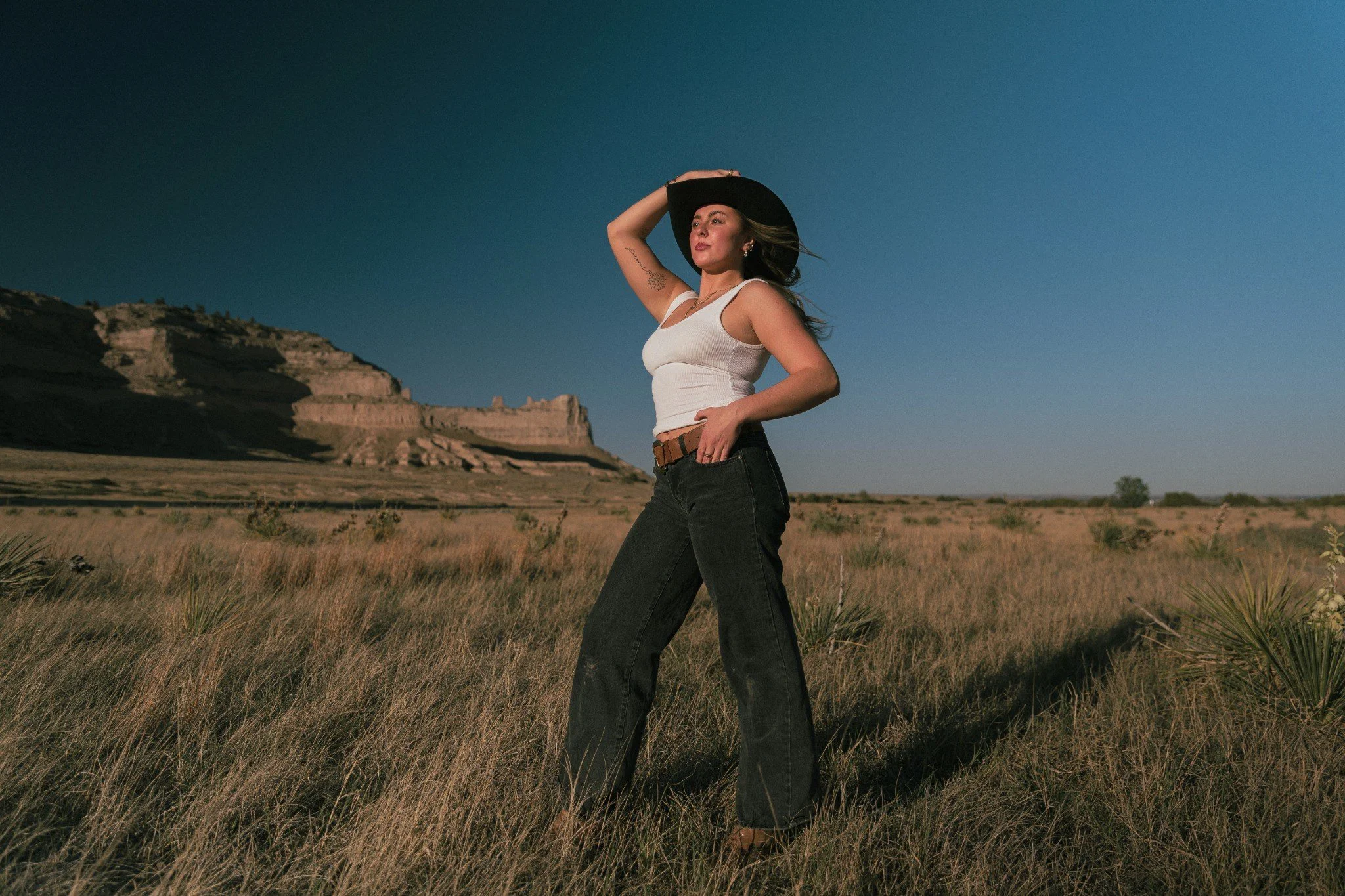Woman standing in a field with mountains in the background, wearing a white tank top, black wide-brim hat, and black jeans, touching her hat with one hand and looking into the distance.