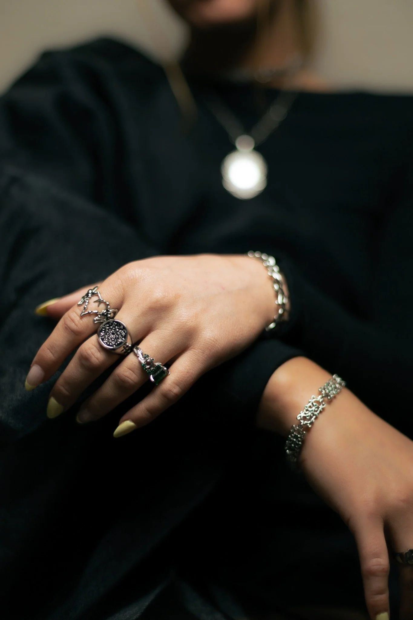 Close-up of a person's hand with multiple rings, wearing yellow nail polish, and a silver bracelet. The person is also wearing a black long-sleeve shirt with part of the hand pointing downward.