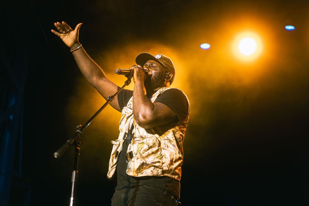 A male singer performing on stage with a microphone, raising one hand, wearing a cap, glasses, a black t-shirt, and a patterned vest, with stage lights illuminating him.