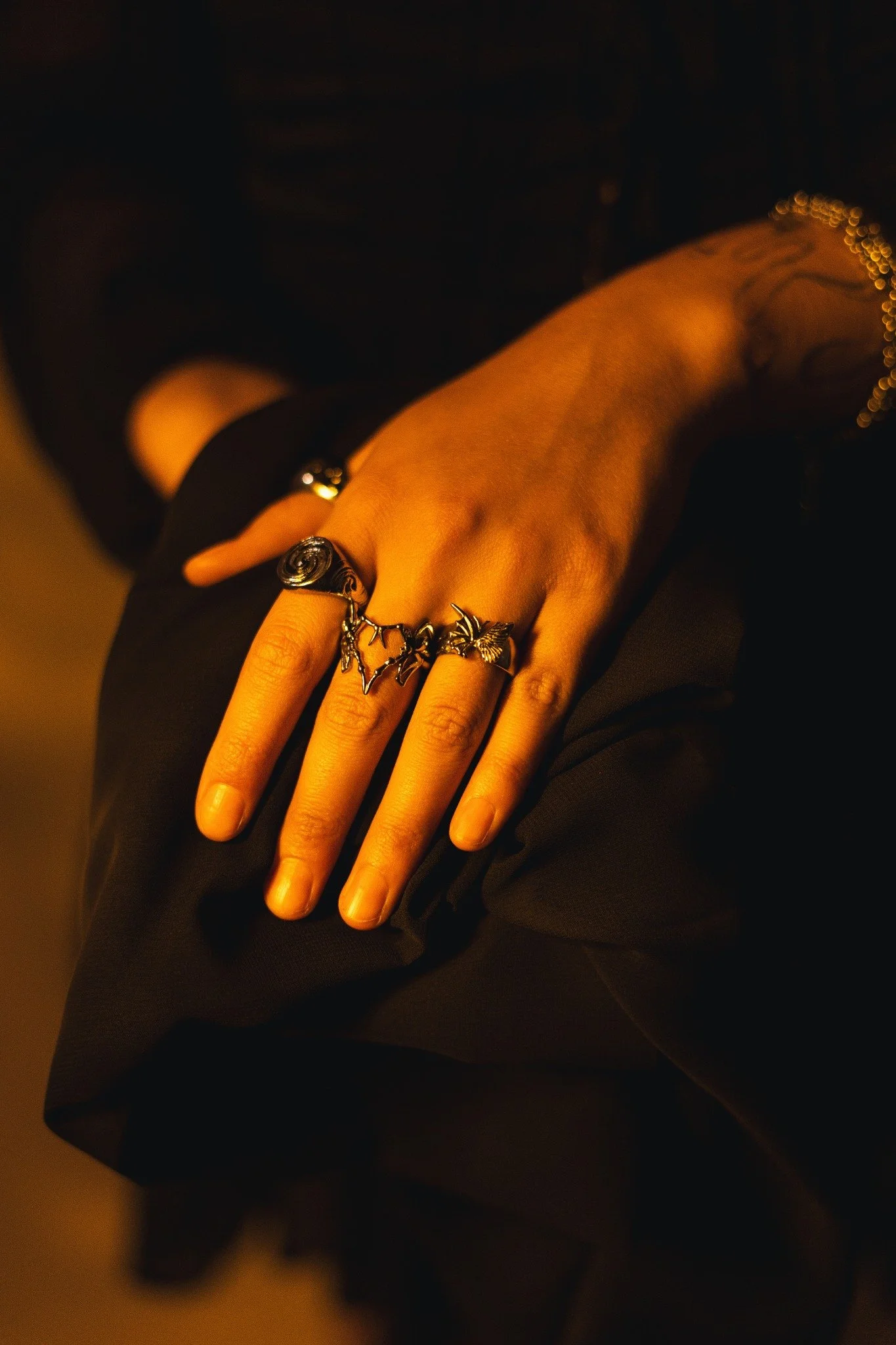 Close-up of a person's hand wearing several rings, resting on their lap, with dark clothing and dim lighting.