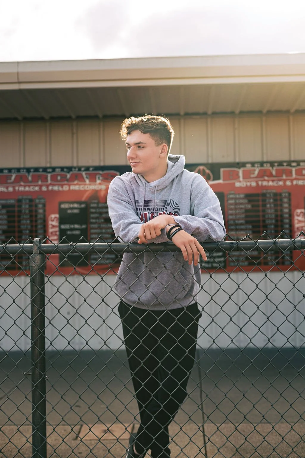 A teenage boy in a gray hoodie standing behind a chain-link fence at a sports stadium, with a red banners and scoreboard in the background.