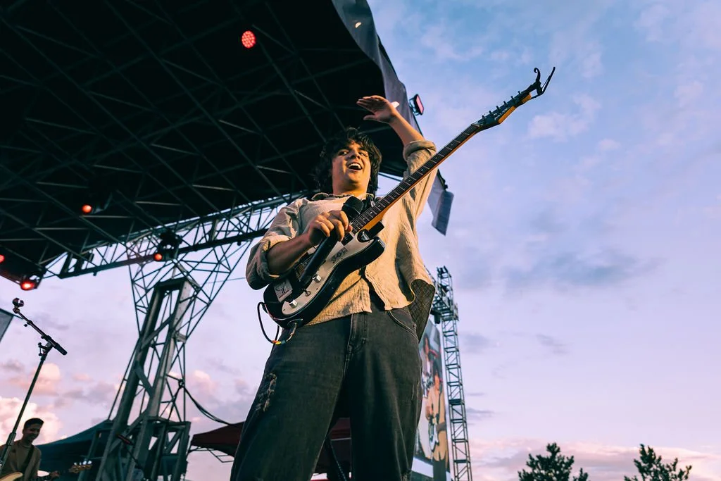 A young man on an outdoor stage playing an electric guitar and waving his hand, with a sky and trees in the background.