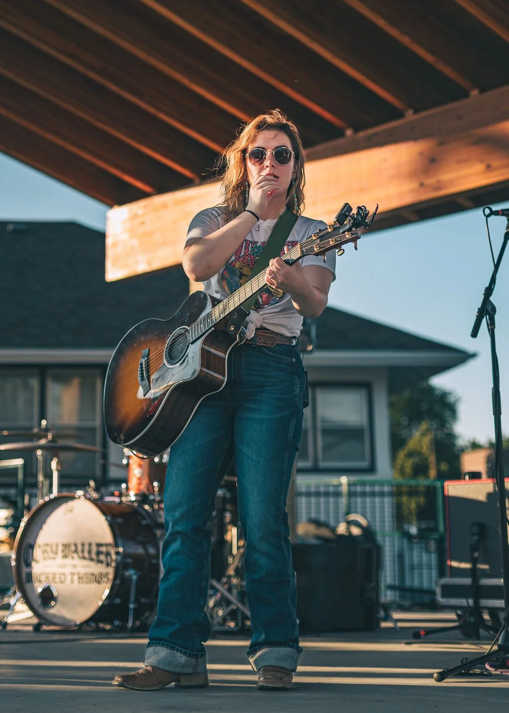 A young woman with wavy hair, wearing sunglasses and casual clothing, is standing on a stage playing an acoustic guitar.