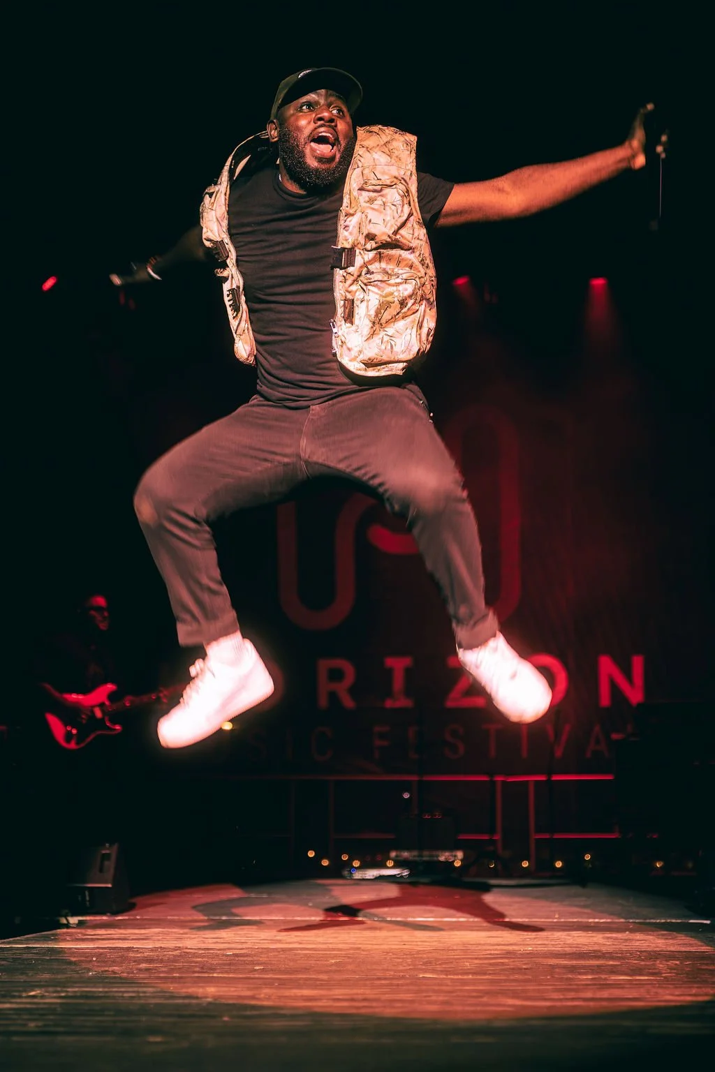 Performer jumping in the air on stage at the Arizona Music Festival, holding a microphone, with a guitar player in the background.