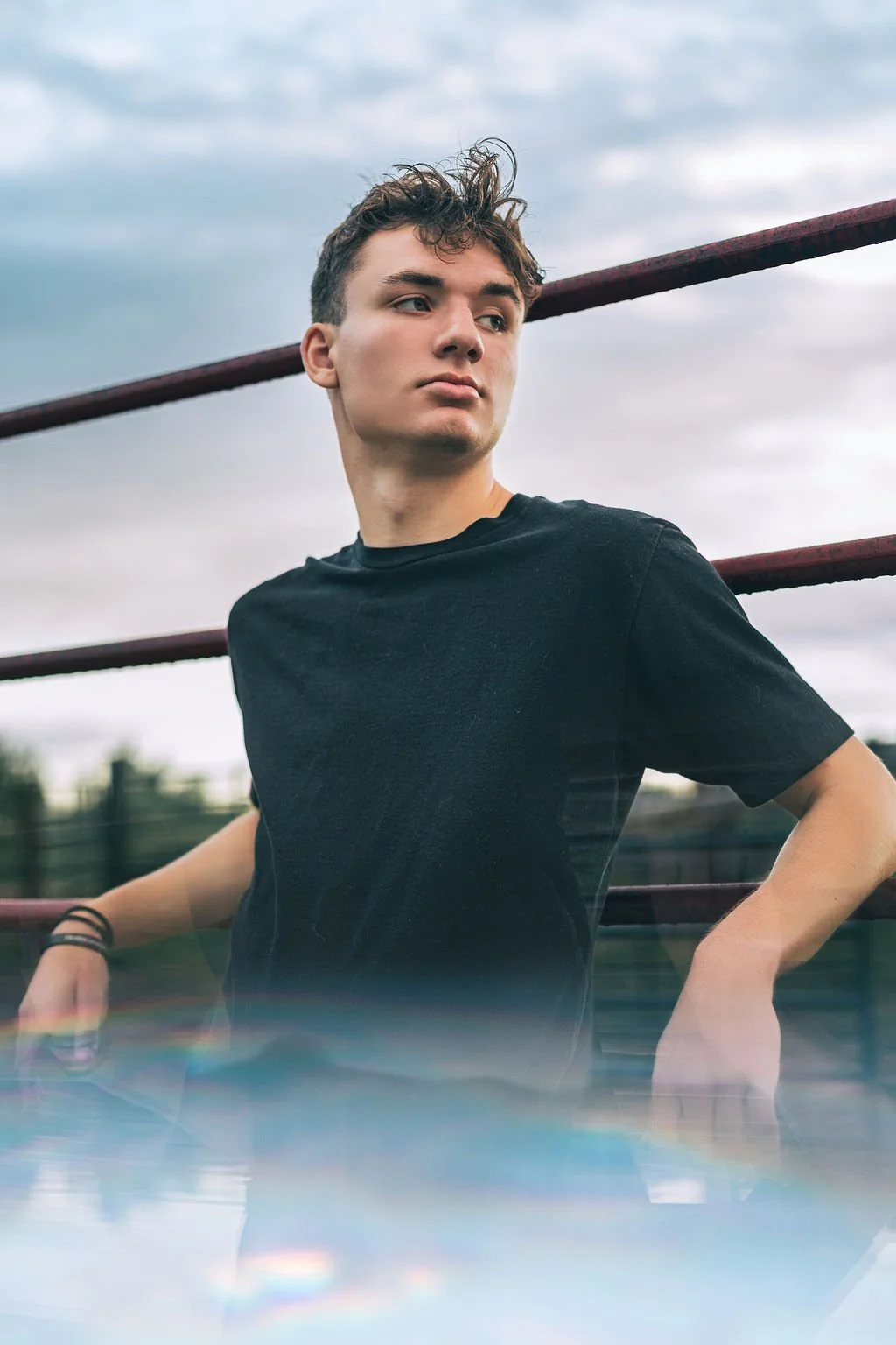 Young man with curly hair wearing a black t-shirt, standing outdoors near a red railing, with a cloudy sky background.
