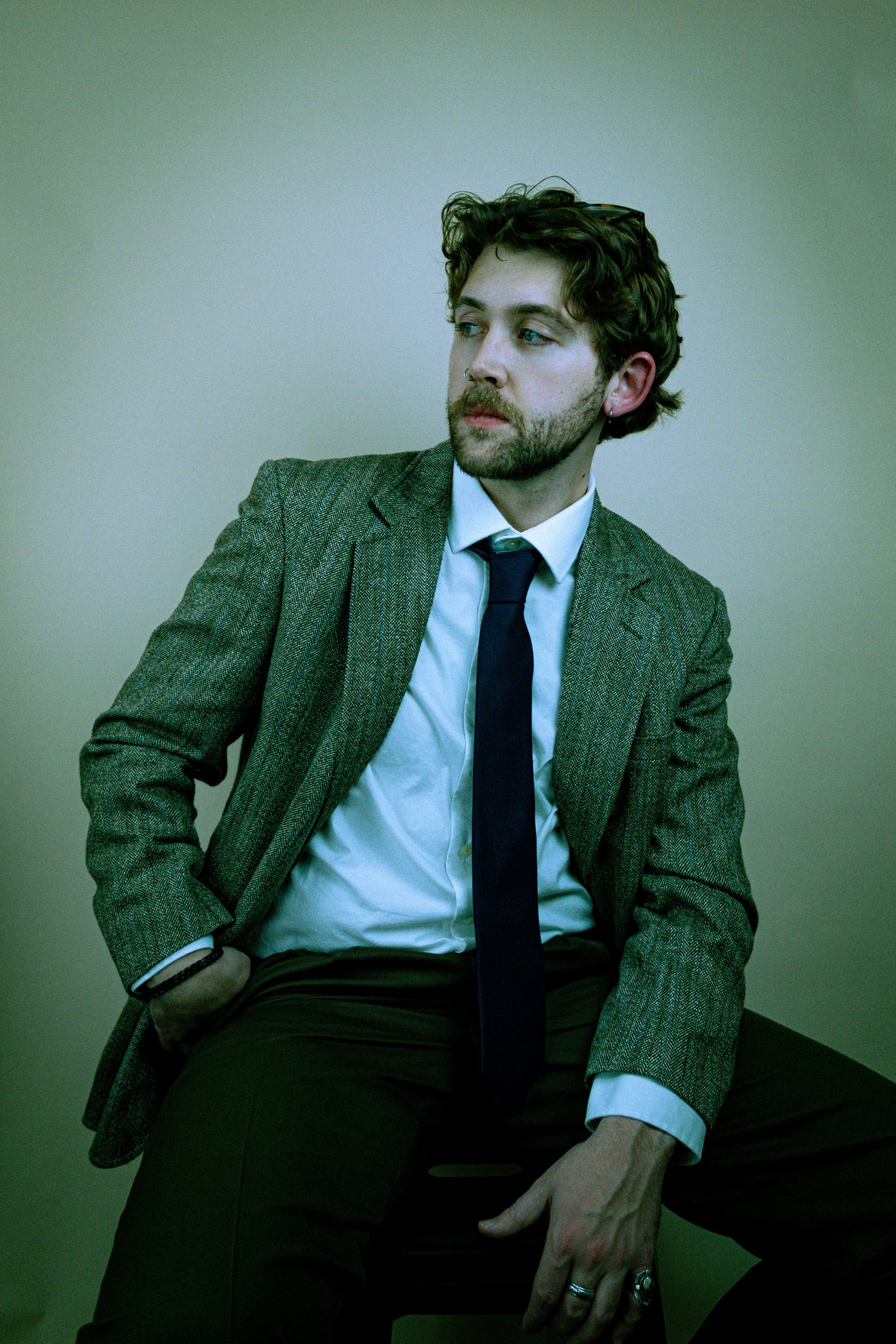A young man with curly hair, a beard, and a nose ring, dressed in a white shirt, dark tie, and grey plaid blazer, sits on a black chair against a plain wall. He has sunglasses resting on his head and wears rings and bracelets.