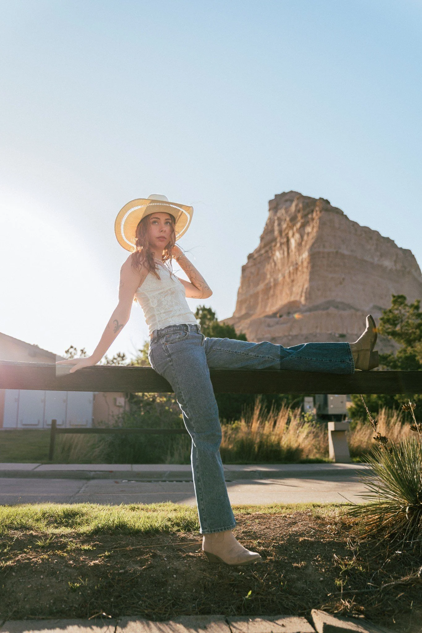 A young woman with wavy hair, wearing a wide-brimmed hat, sleeveless top, jeans, and boots, sitting on a ledge in front of a rock formation during sunset.