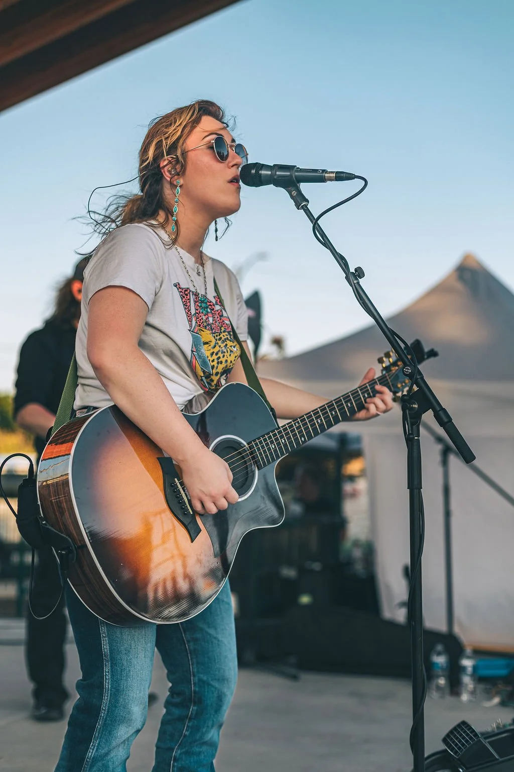 A female musician with wavy blonde hair, wearing sunglasses and earrings, singing into a microphone while playing an acoustic guitar during an outdoor performance at sunset.