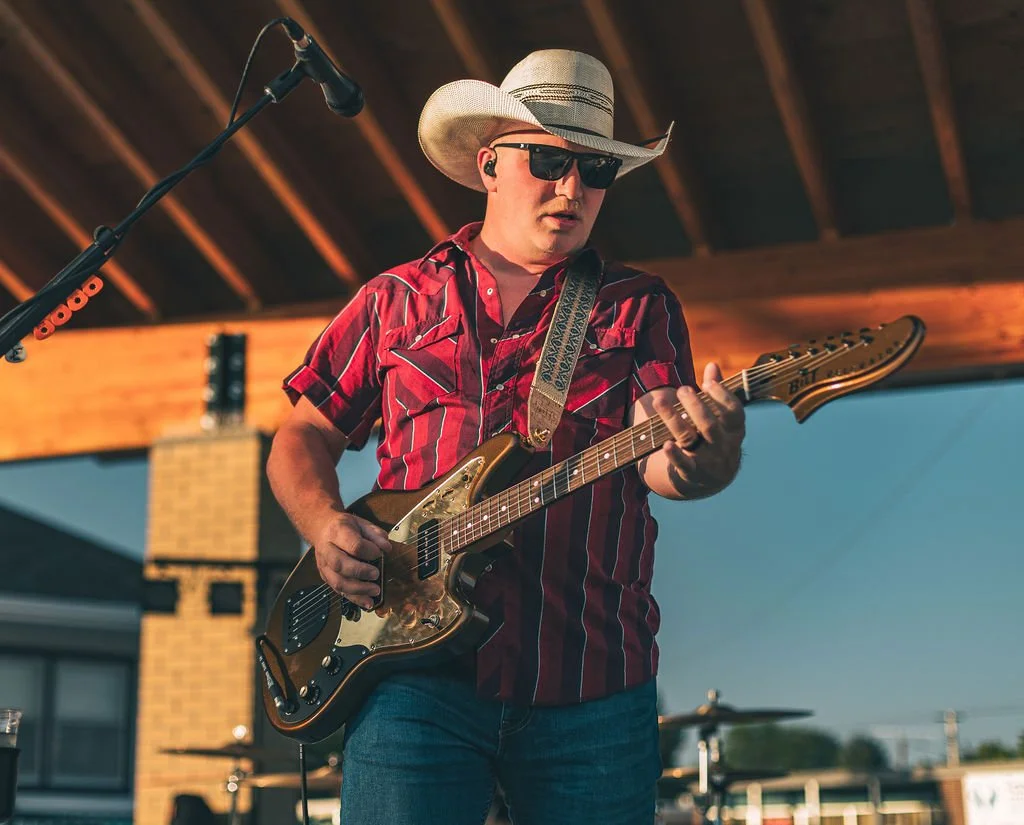 A man wearing a cowboy hat, sunglasses, and a red striped shirt playing an electric guitar on a stage outdoors.