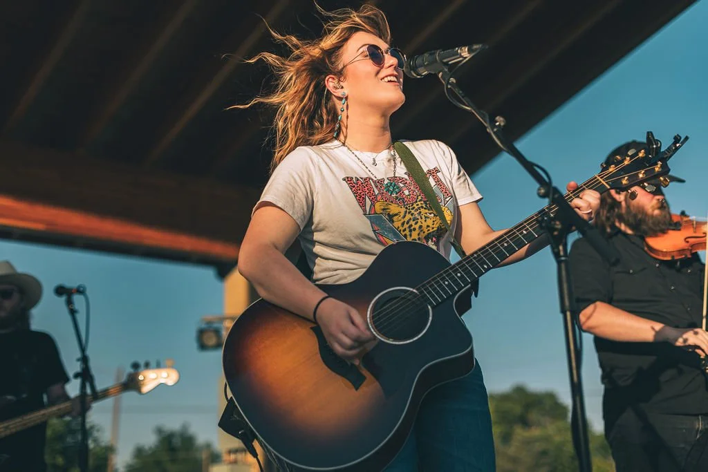 Young woman with sunglasses playing an acoustic guitar and singing into a microphone during an outdoor performance, with other musicians in the background.