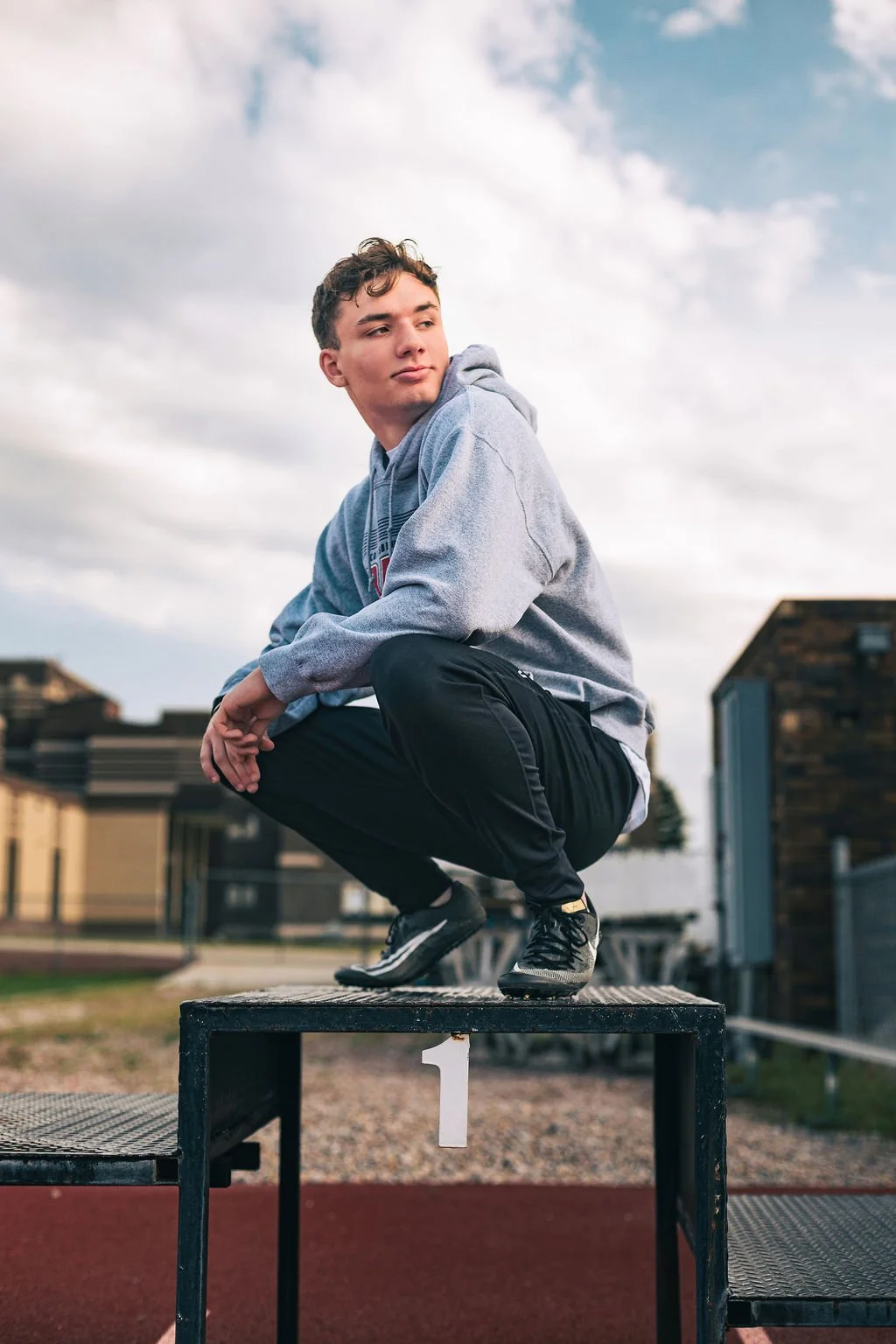 Young man squatting on a black podium marked with a white number '1' at an outdoor sports field, wearing a gray hoodie, black pants, and black sneakers, with buildings and cloudy sky in the background.