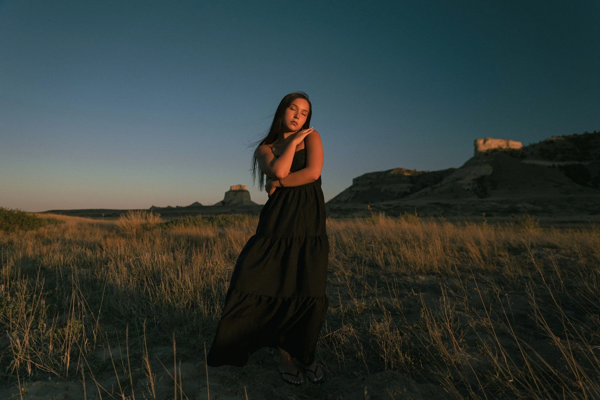 A woman in a black dress standing in a grassy field during sunset with a clear blue sky and rock formations in the background.