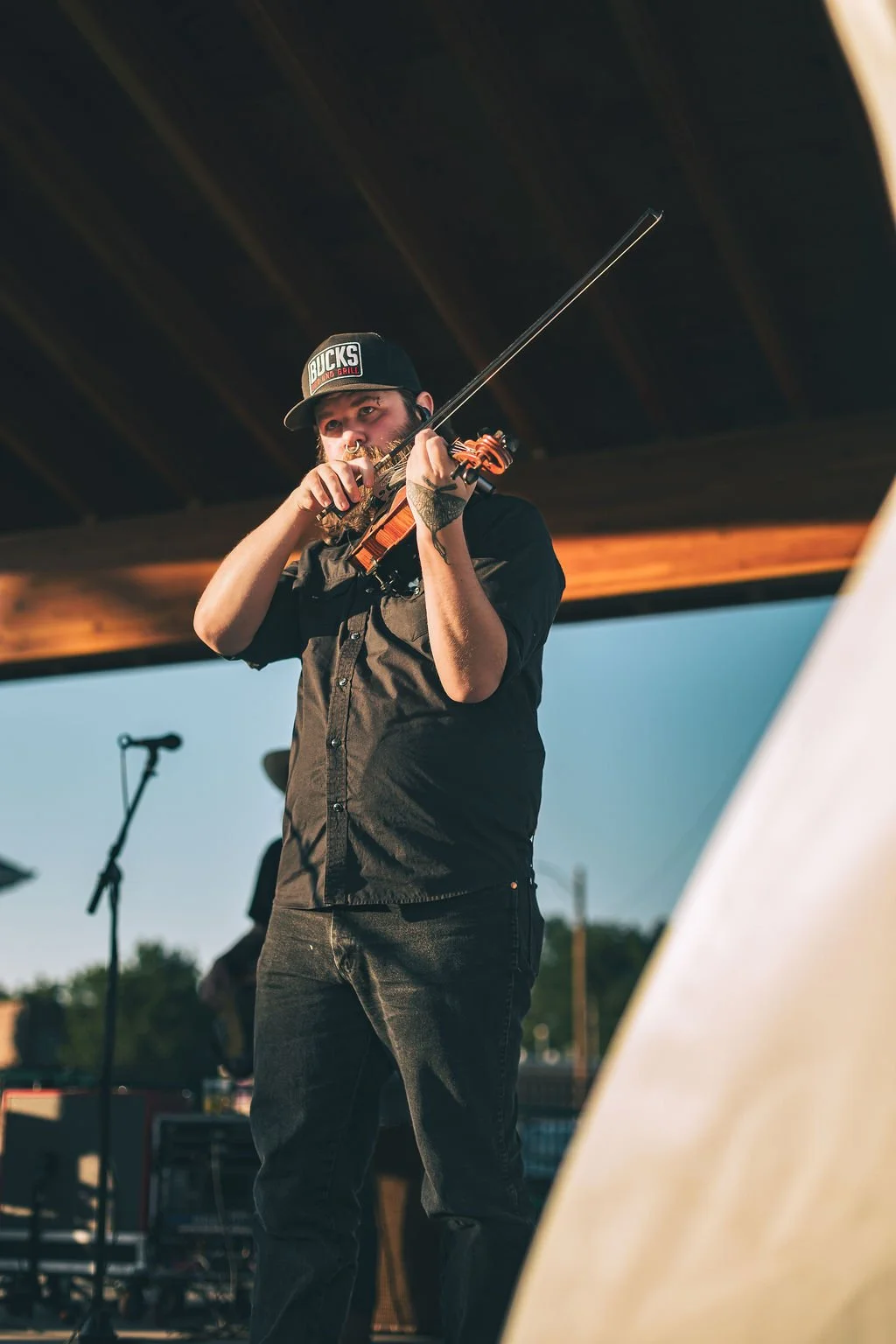 A man playing a violin on an outdoor stage during sunset, wearing a black shirt, black jeans, and a black cap with red and white text, with a microphone and musical equipment in the background.