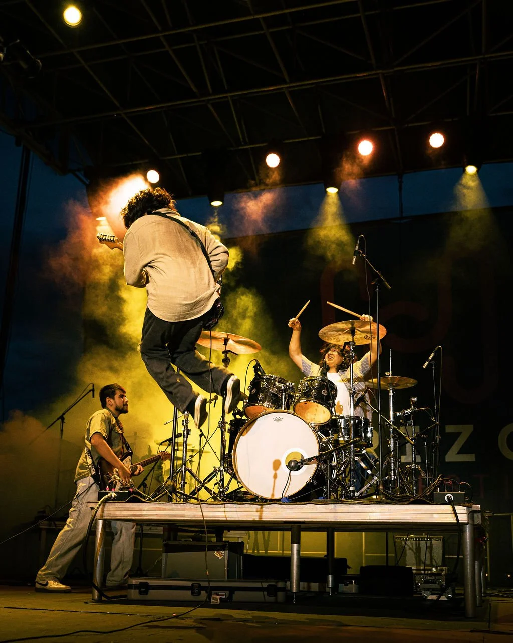 A live band performance on stage with one guitarist jumping in the air, a drummer enthusiastically playing, and another musician standing on the side playing a guitar, under colorful stage lights.