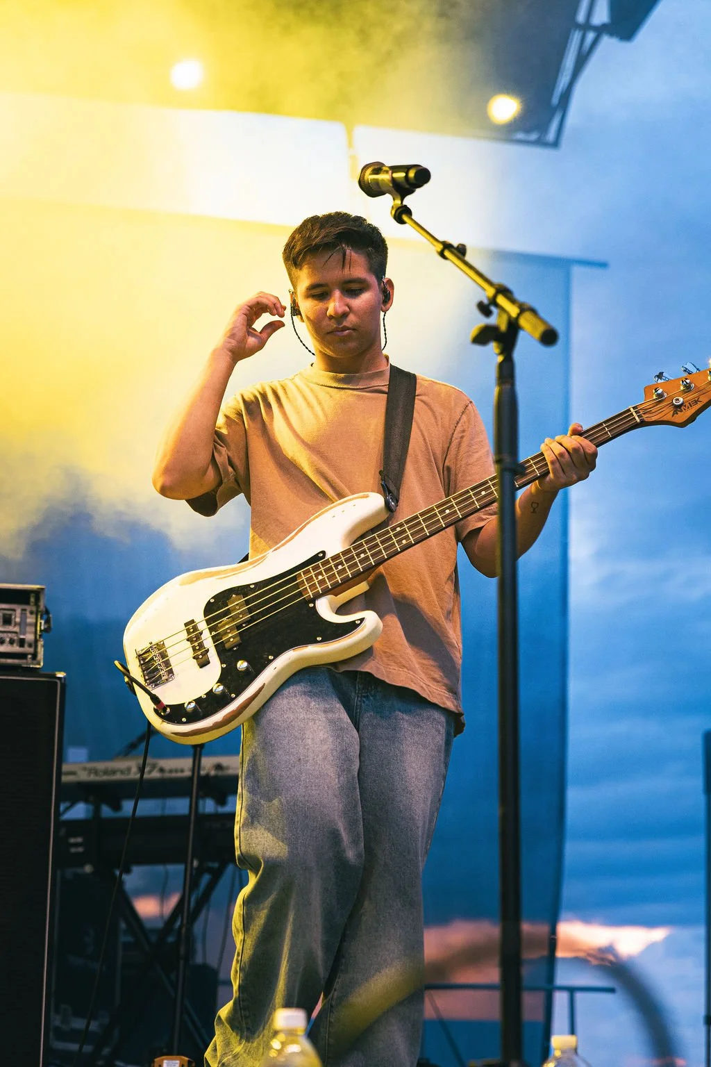 A young male musician with short dark hair wearing a beige T-shirt and blue jeans, playing a white bass guitar on stage with yellow and blue lighting, a microphone stand, and musical equipment in the background.