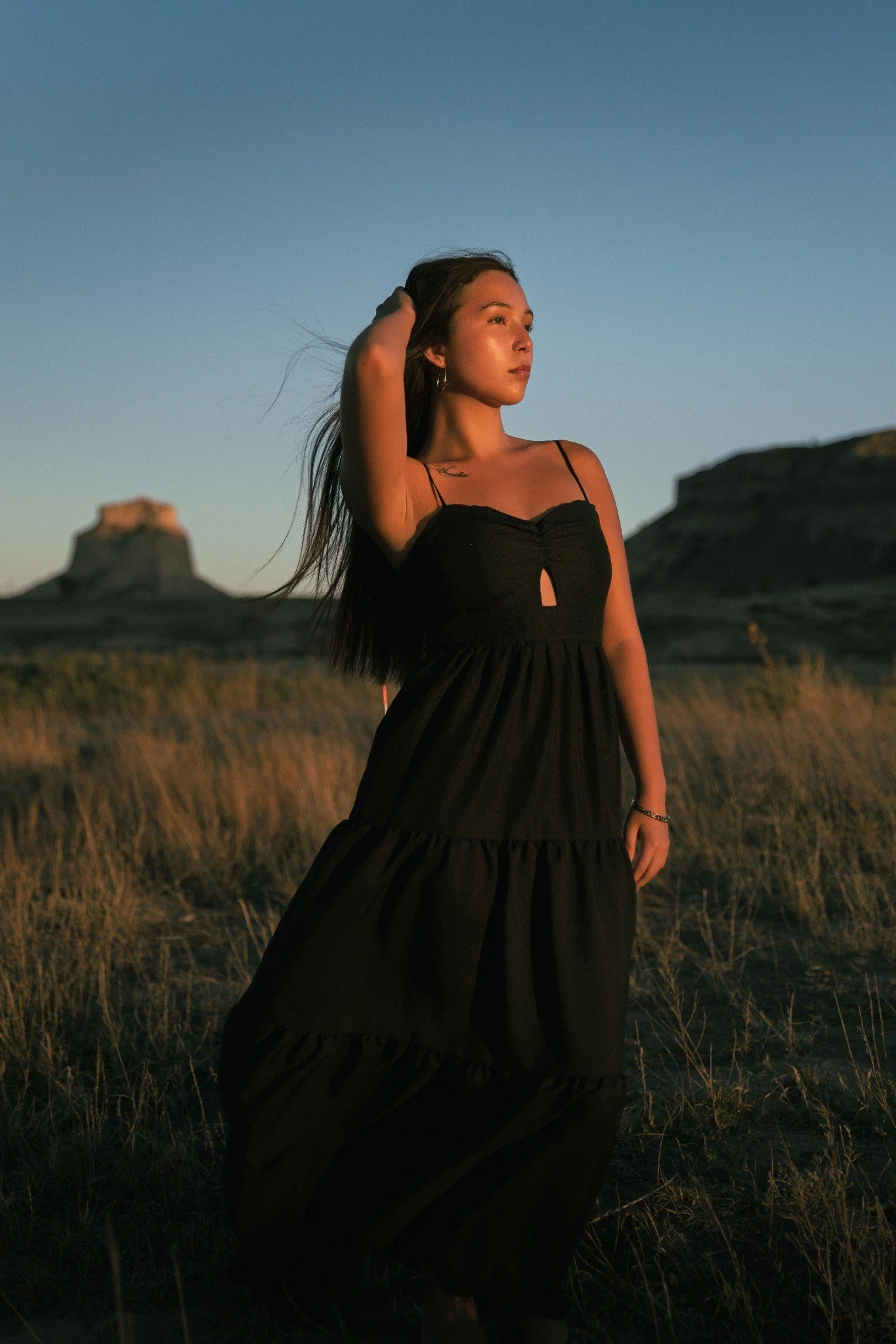 A woman with long hair in a black dress standing in a field at sunset or sunrise, with rock formations in the background.