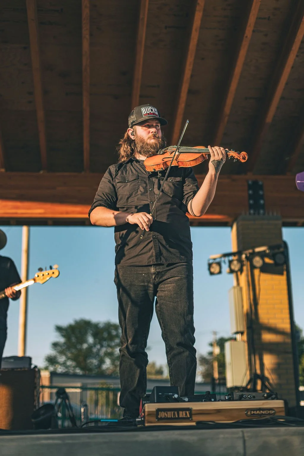 A man with long hair and a beard playing a violin on a stage outdoors, wearing a black shirt and a cap, with a guitarist visible in the background and stage equipment nearby.