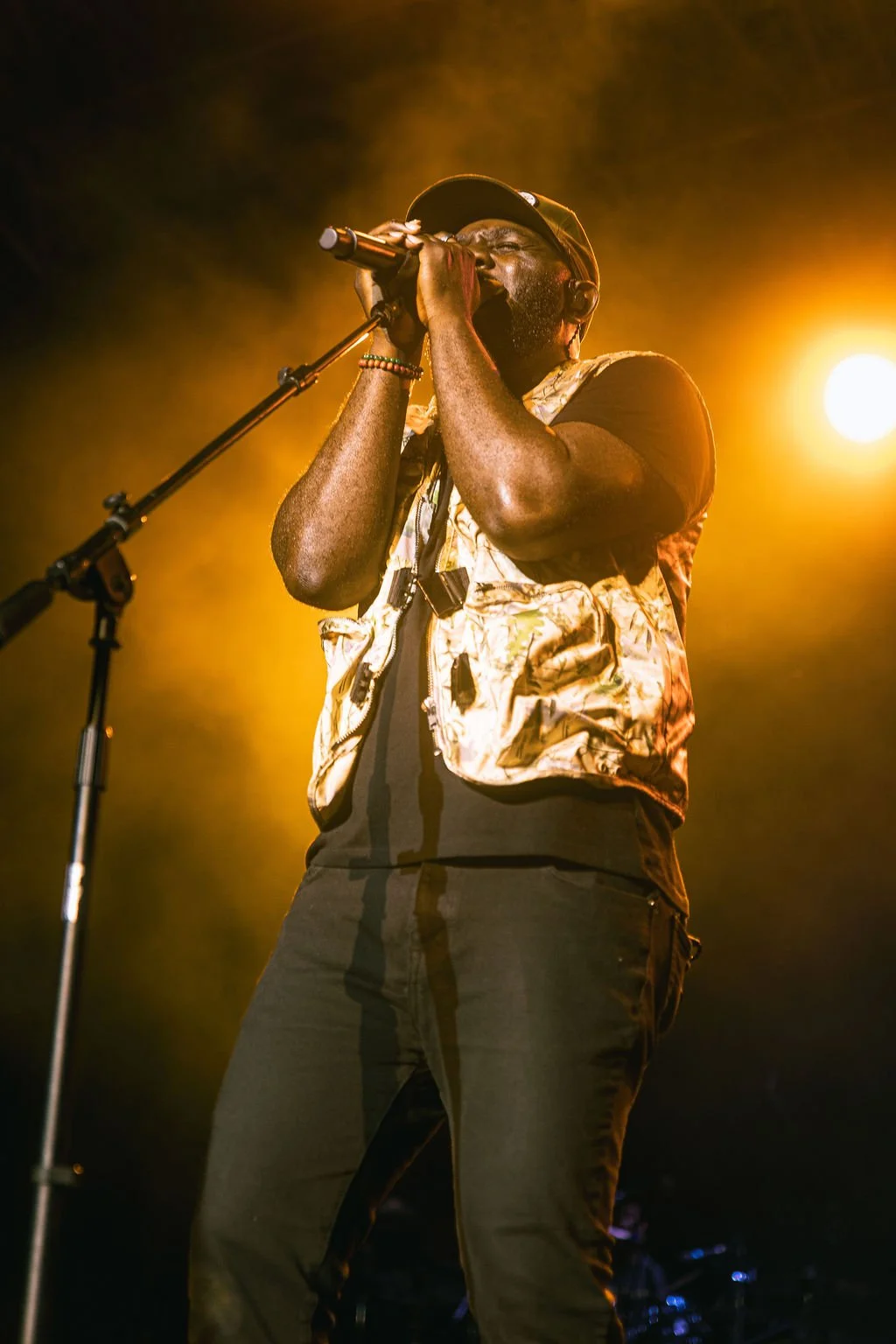 A man singing into a microphone on stage, illuminated by yellow stage lights, wearing a cap, a patterned vest, and black pants.