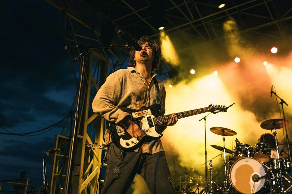 A male guitarist and singer performing on stage during a concert at night, with colorful stage lights and a drum set in the background.