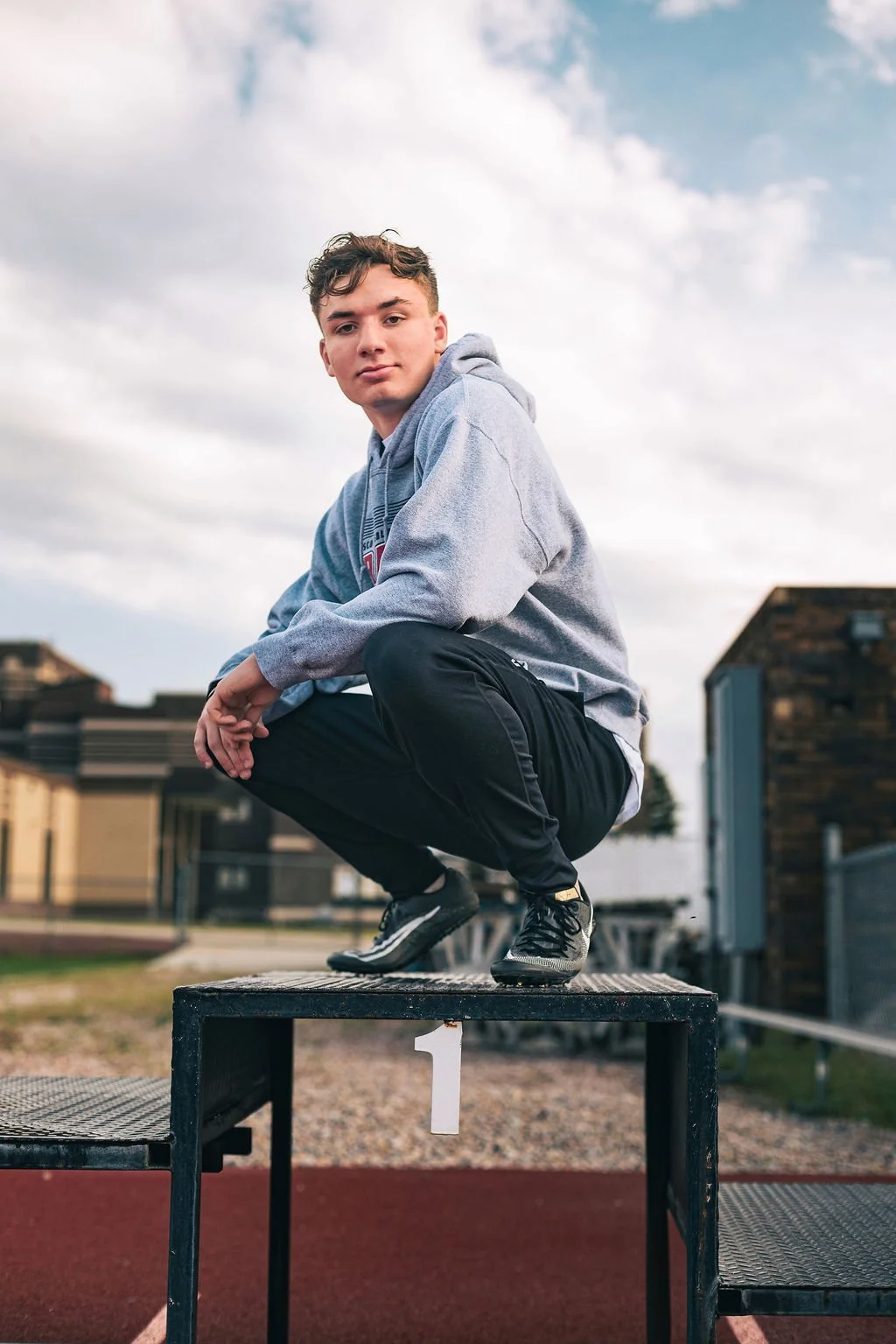 Young man squatting on a winners' pedestal at an outdoor sports track, with a cloudy sky in the background.