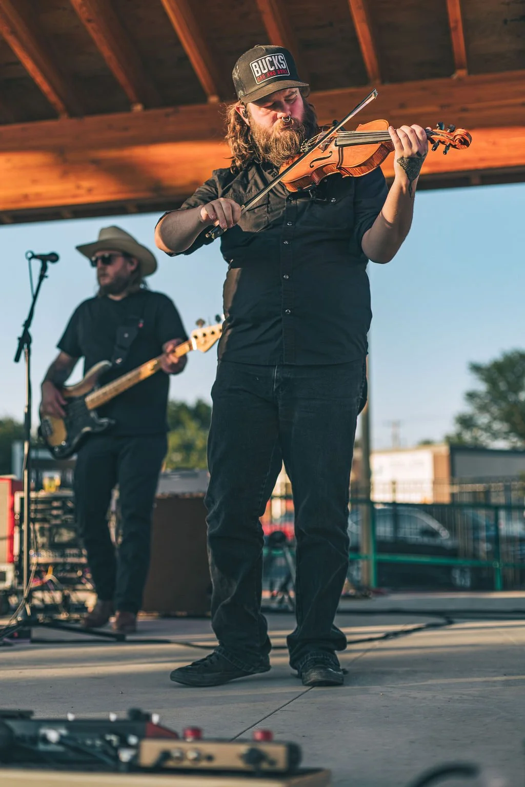 A man with a beard and nose ring playing a violin on an outdoor stage, wearing a black shirt and a baseball cap, with another musician playing an electric bass guitar in the background under a wooden roof.