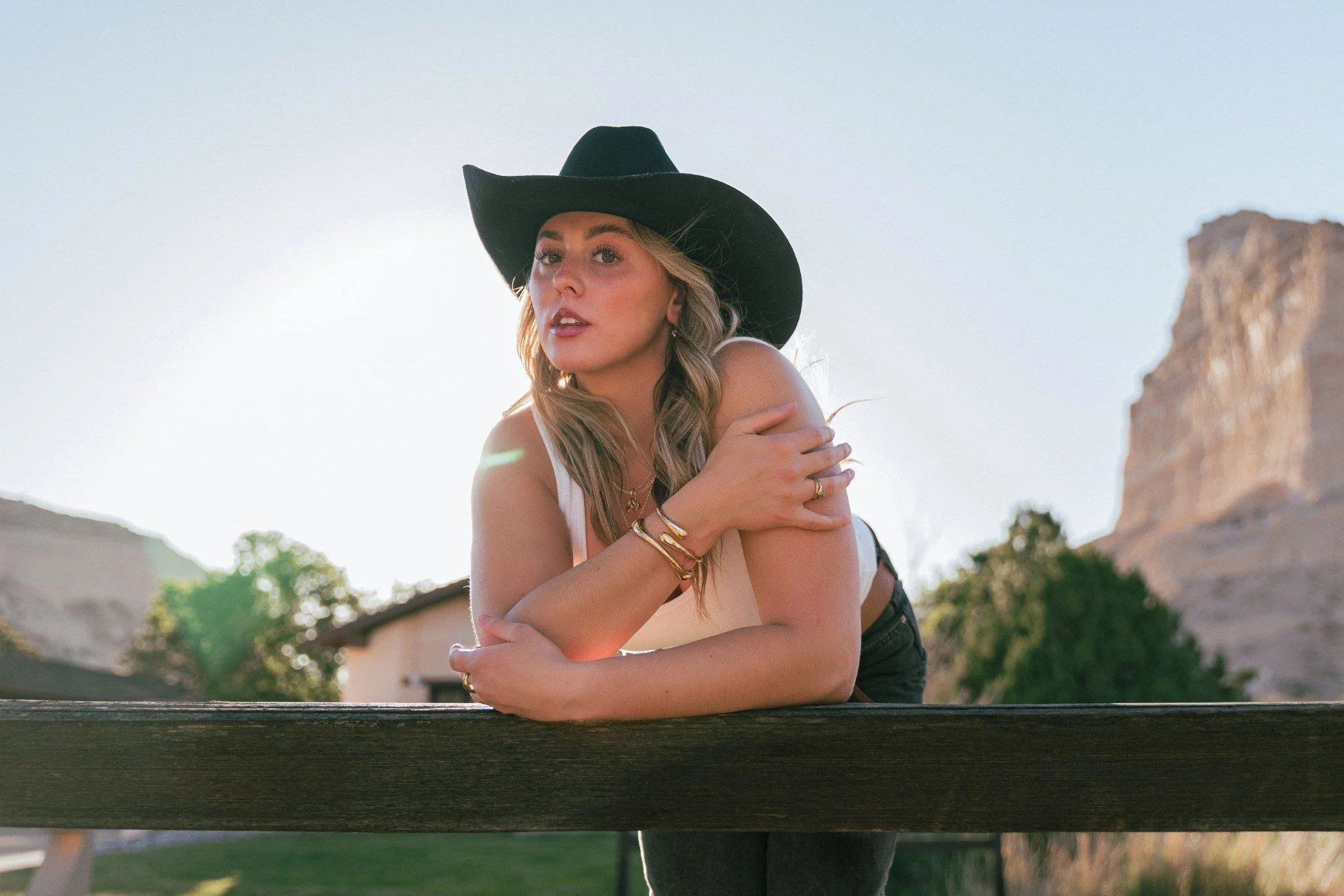 A woman with blonde hair wearing a black cowboy hat and gold jewelry, sitting outdoors during daylight, with rocks and trees in the background.