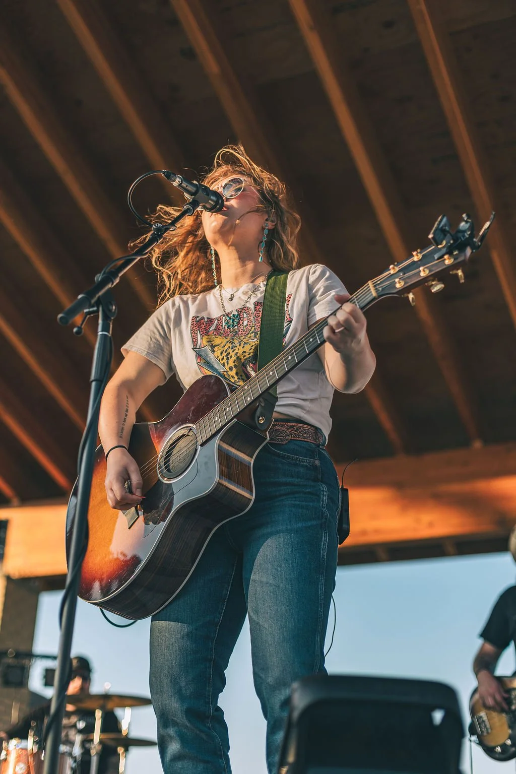 A woman with curly hair and sunglasses singing into a microphone while playing an acoustic guitar on stage during a live outdoor performance.