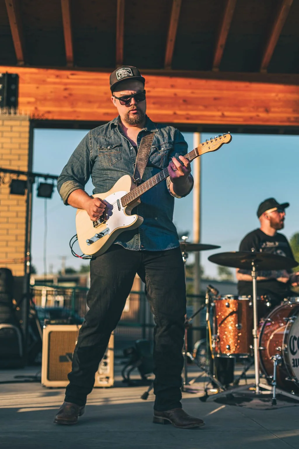 A man wearing sunglasses, a denim jacket, and a cap, playing an electric guitar on an outdoor stage during sunset, with a drummer in the background.