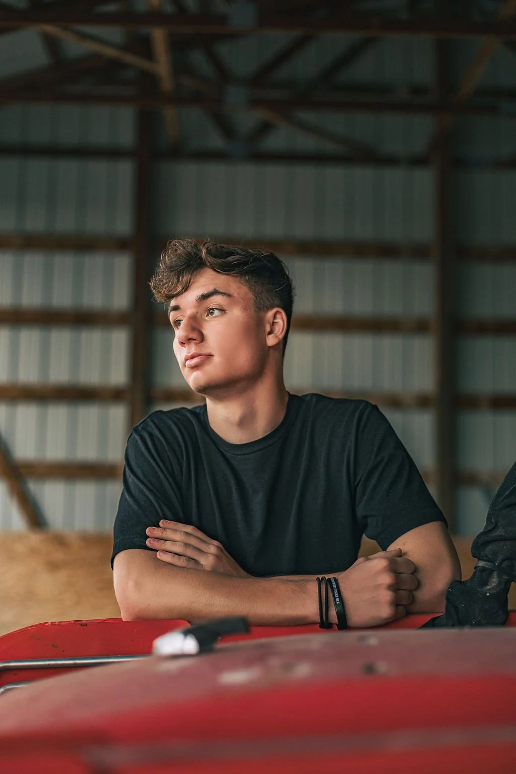 A young man with dark hair, wearing a black t-shirt, sits at a table with his arms crossed inside a structure with wooden beams and metal walls.