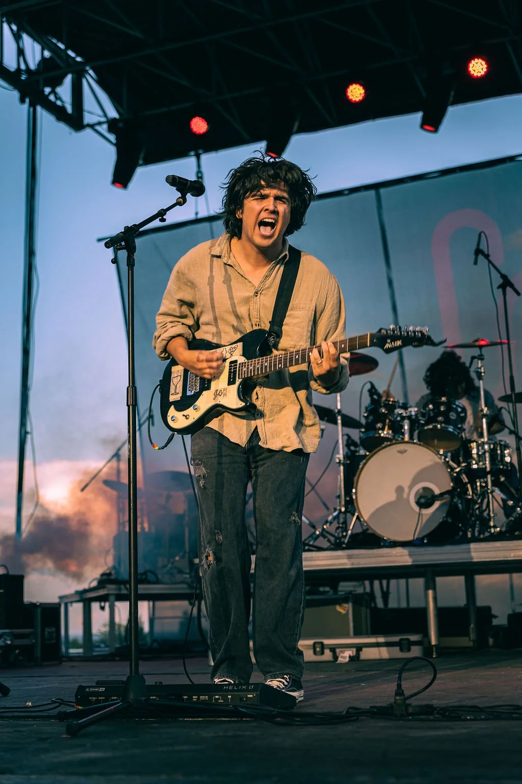 A young male guitarist with messy hair passionately performing on stage, singing into a microphone with a drummer in the background during a live outdoor concert.