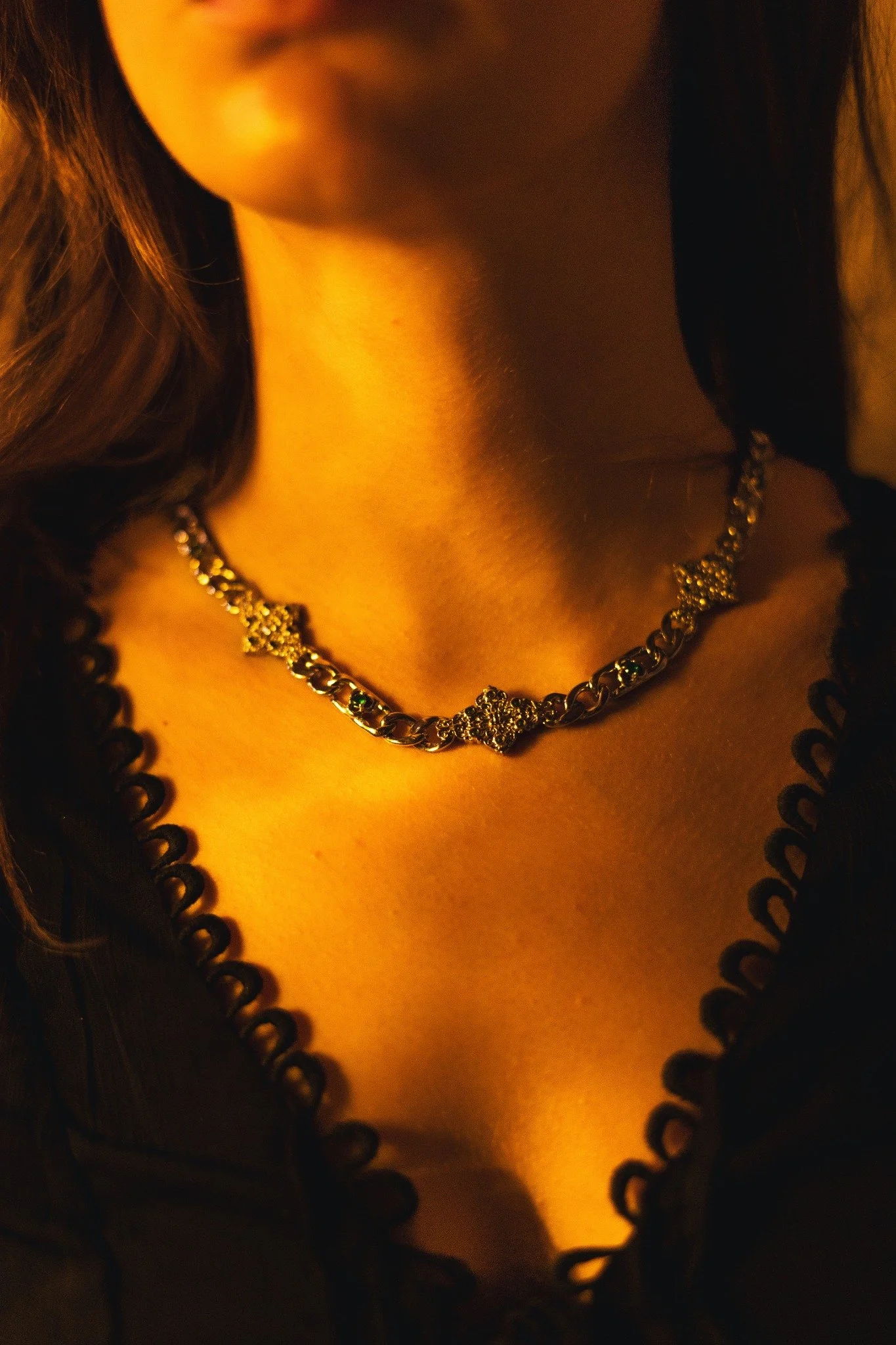 Close-up of a woman's neck and shoulder, wearing a gold necklace with a floral design, in a warm ambient light.
