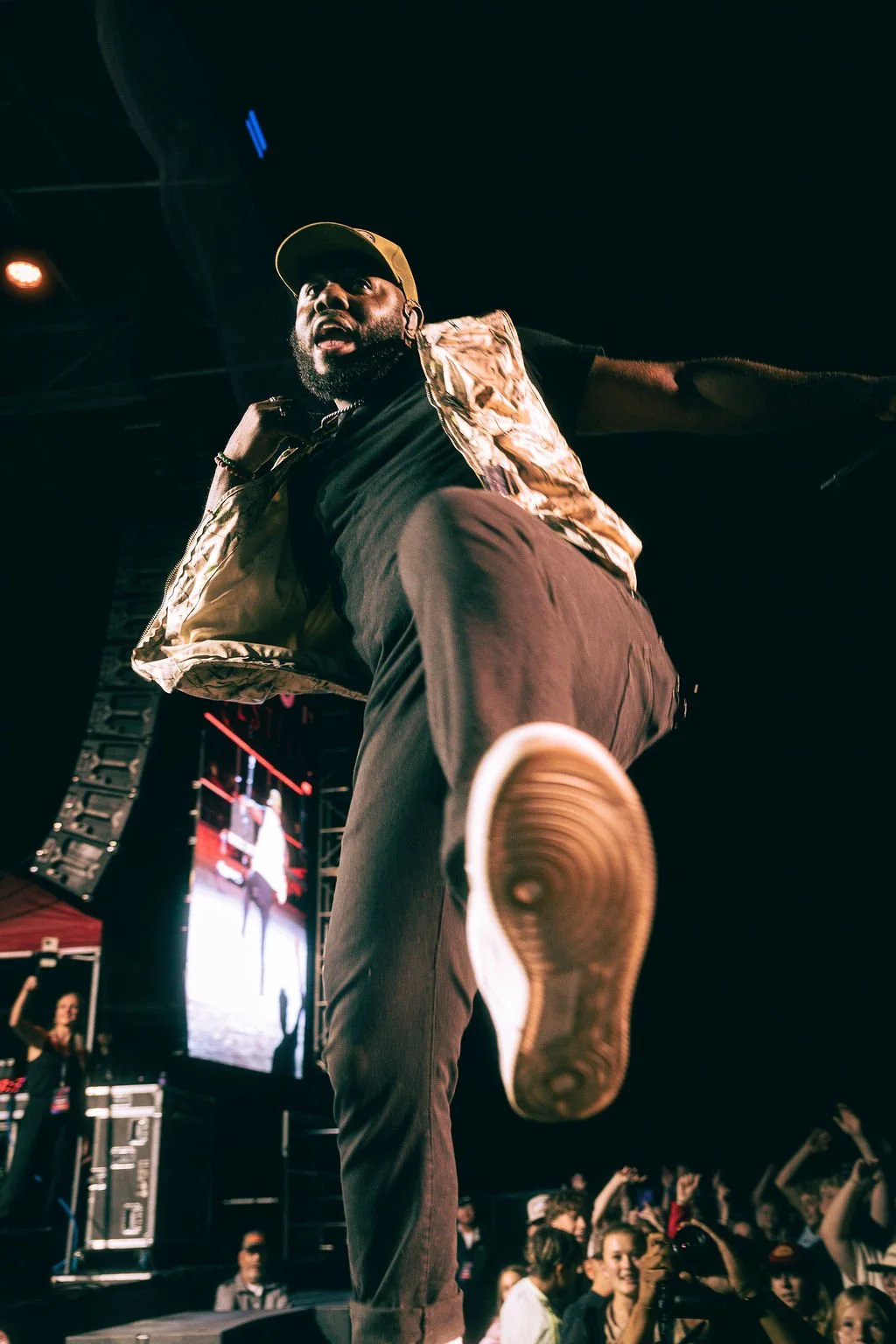 A performer on stage, kicking towards the camera, with an audience and a large screen in the background.