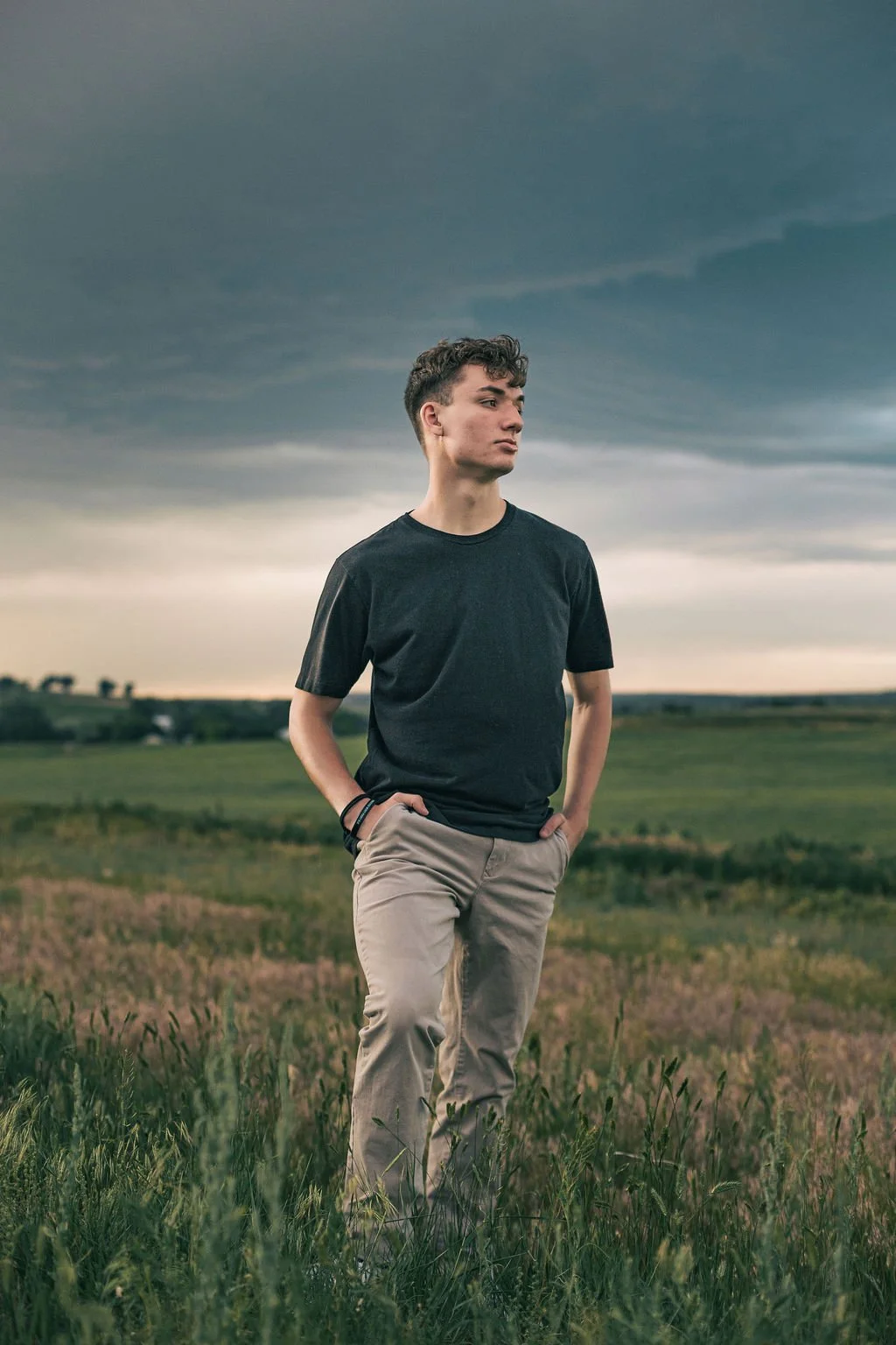 A young man standing in a grassy field under a cloudy sky, wearing a black t-shirt and beige pants, looking to the side with hands in pockets.