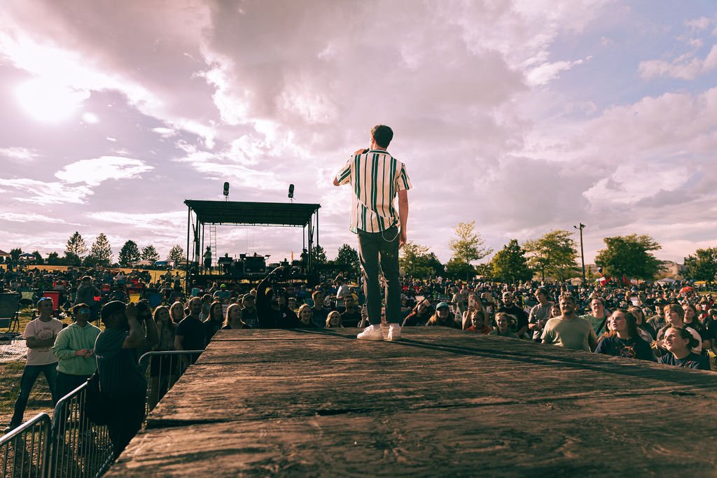 A man in a striped shirt stands on a stage addressing a large outdoor audience during a festival or concert, with a cloudy sky overhead.