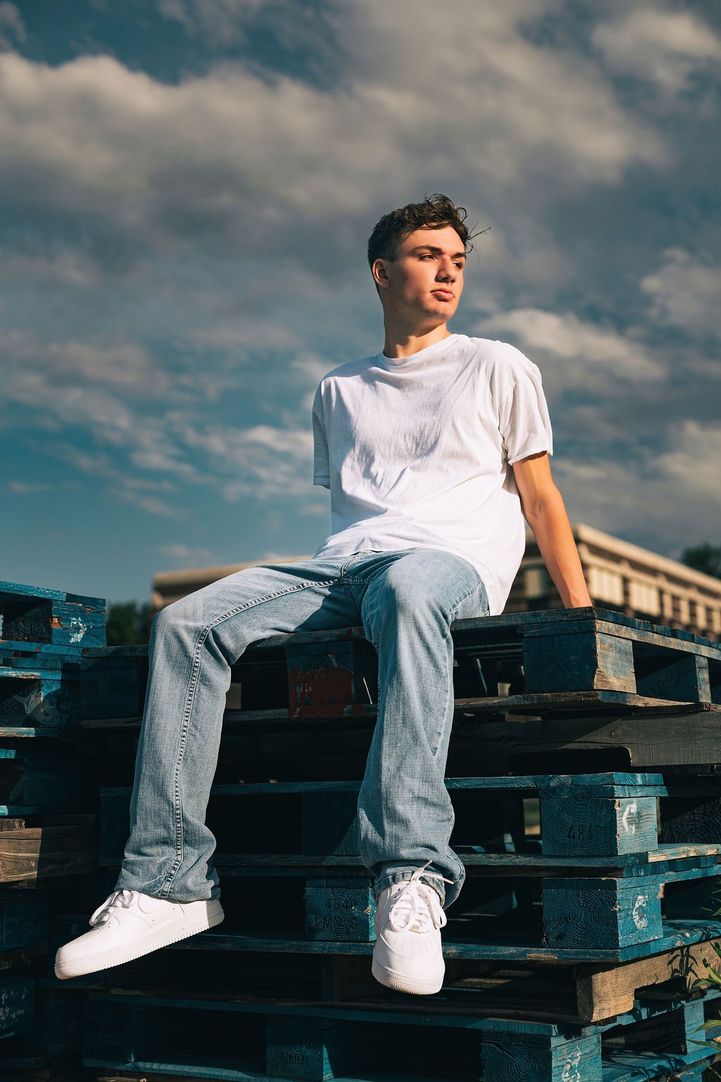 Young man sitting on wooden pallets outdoors, wearing a white t-shirt and light jeans, with a cloudy sky in the background.